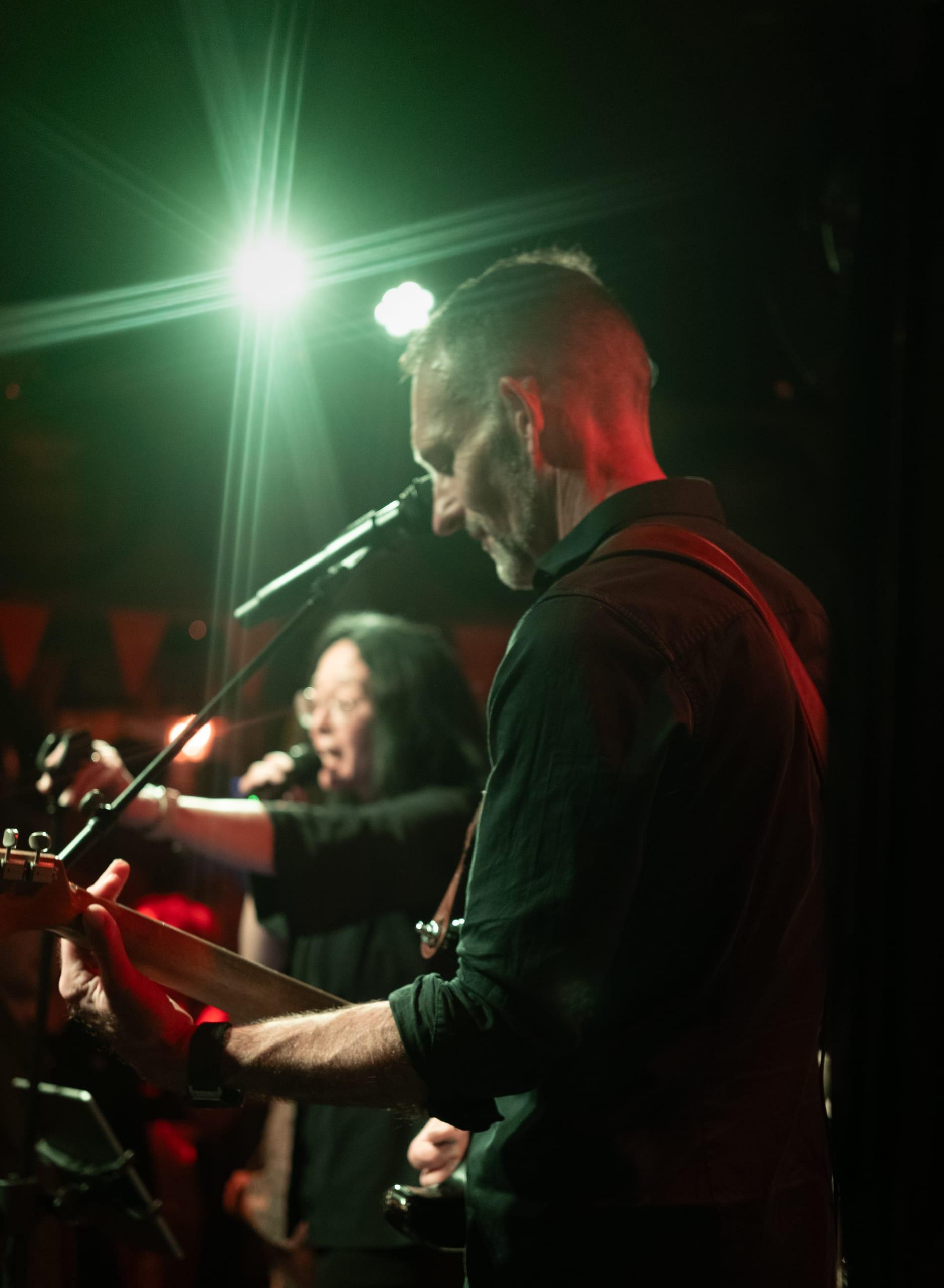 Bassist singing into a mic, second vocalist blurred in the background under green and red stage light