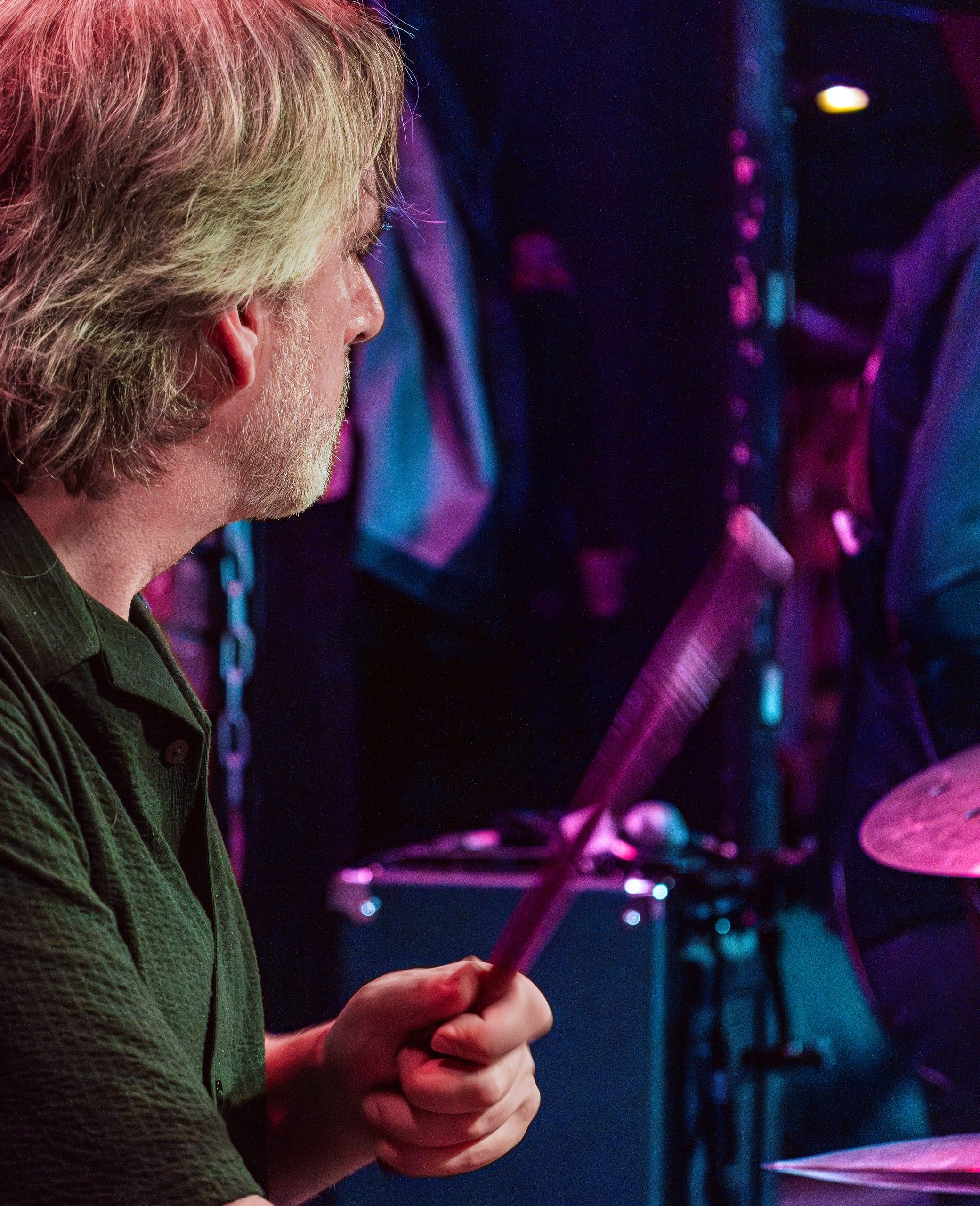 Drummer in profile, sticks crossed mid-strike, lit in pink and blue