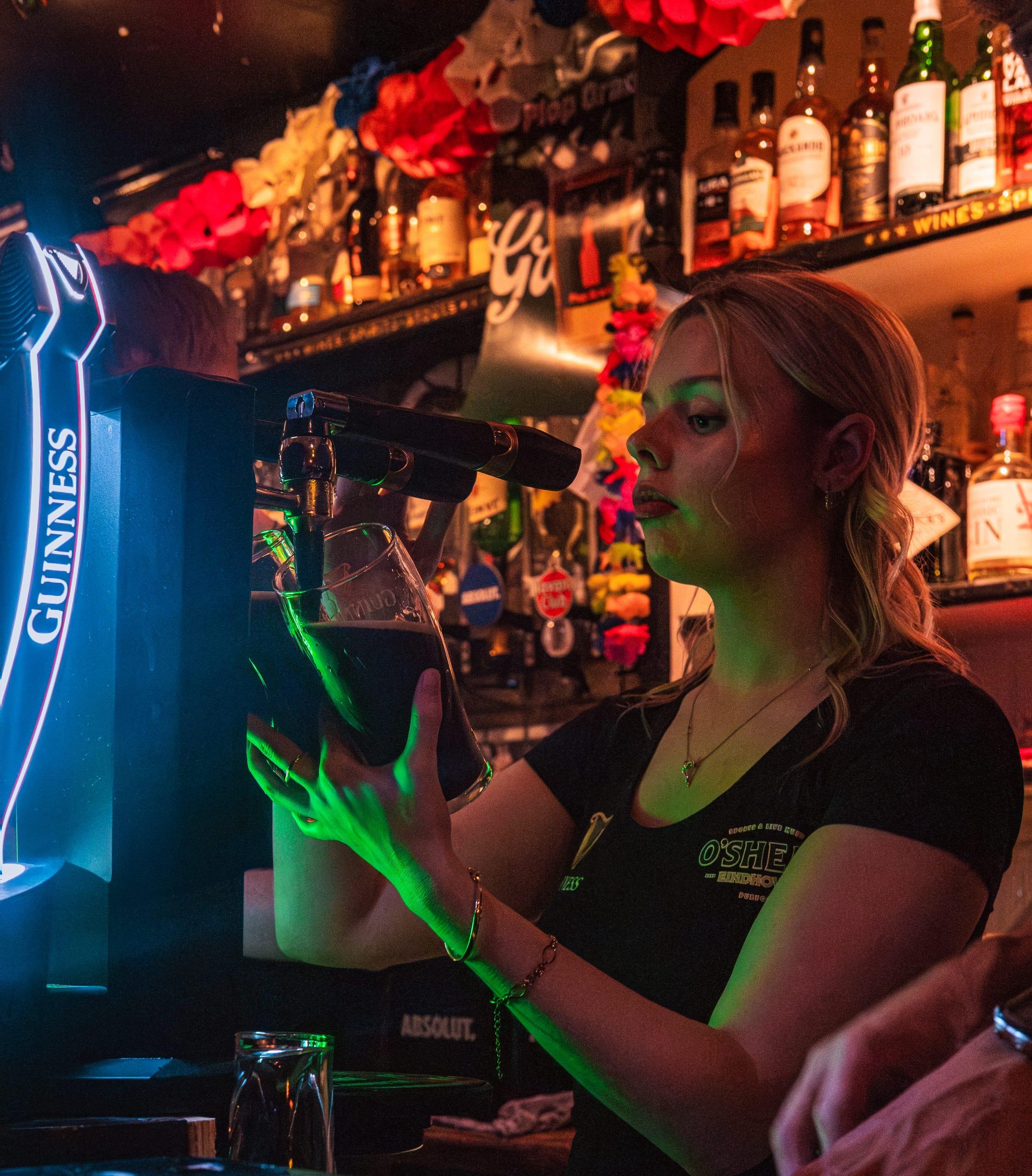 Bartender pulling a pint under green neon Guinness light, O'Shea's shirt visible