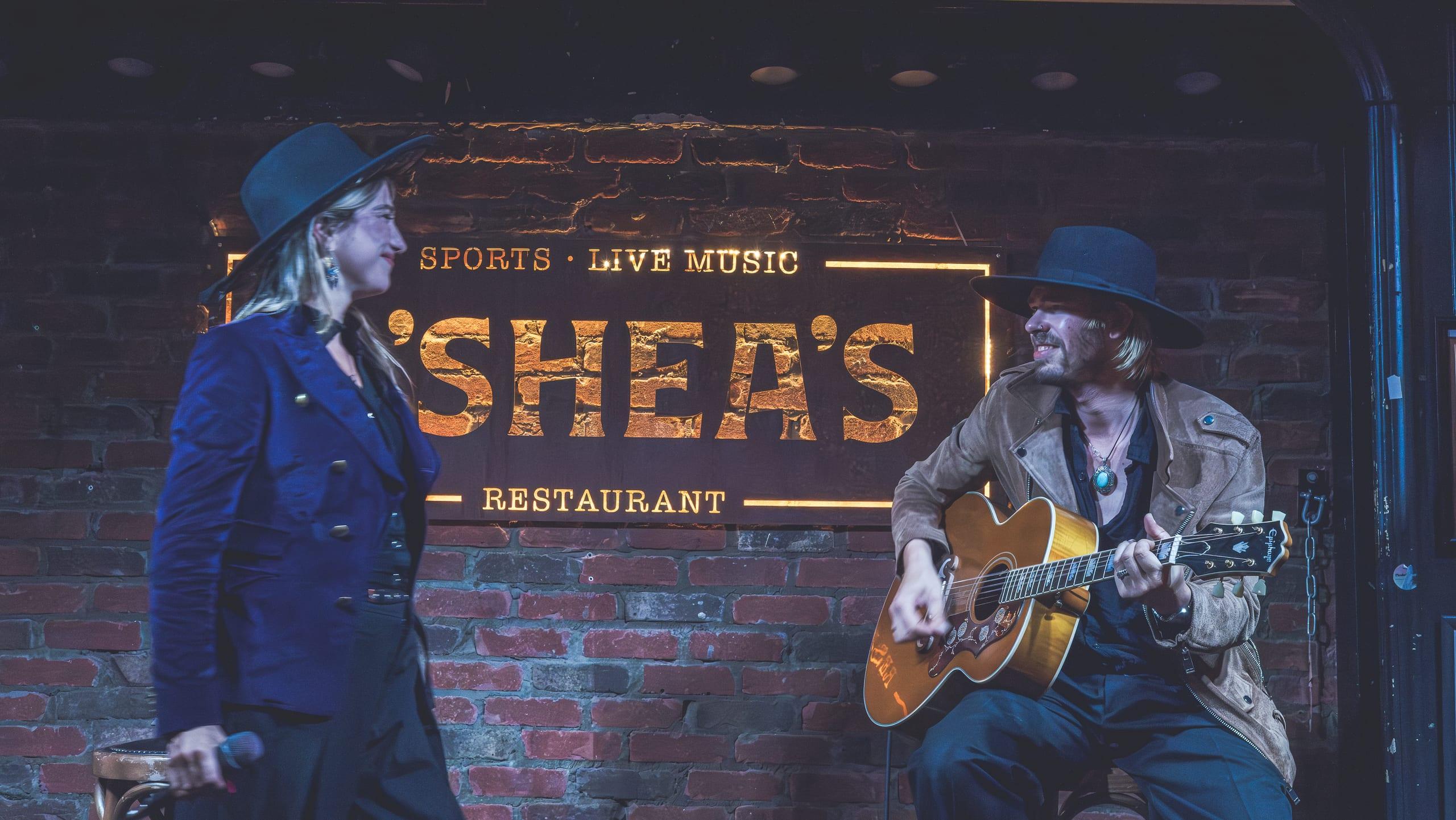 Singer and guitarist trading a look on stage at O'Shea's, full venue sign lit behind them