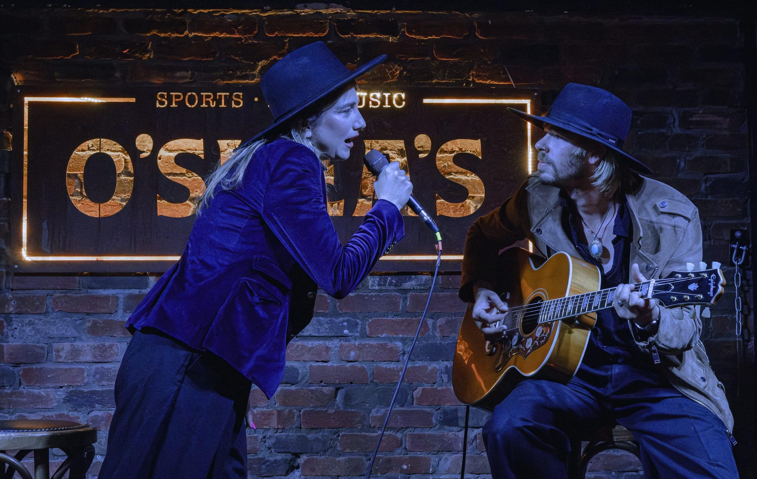 Singer and guitarist facing each other in profile, O'Shea's sign glowing behind them