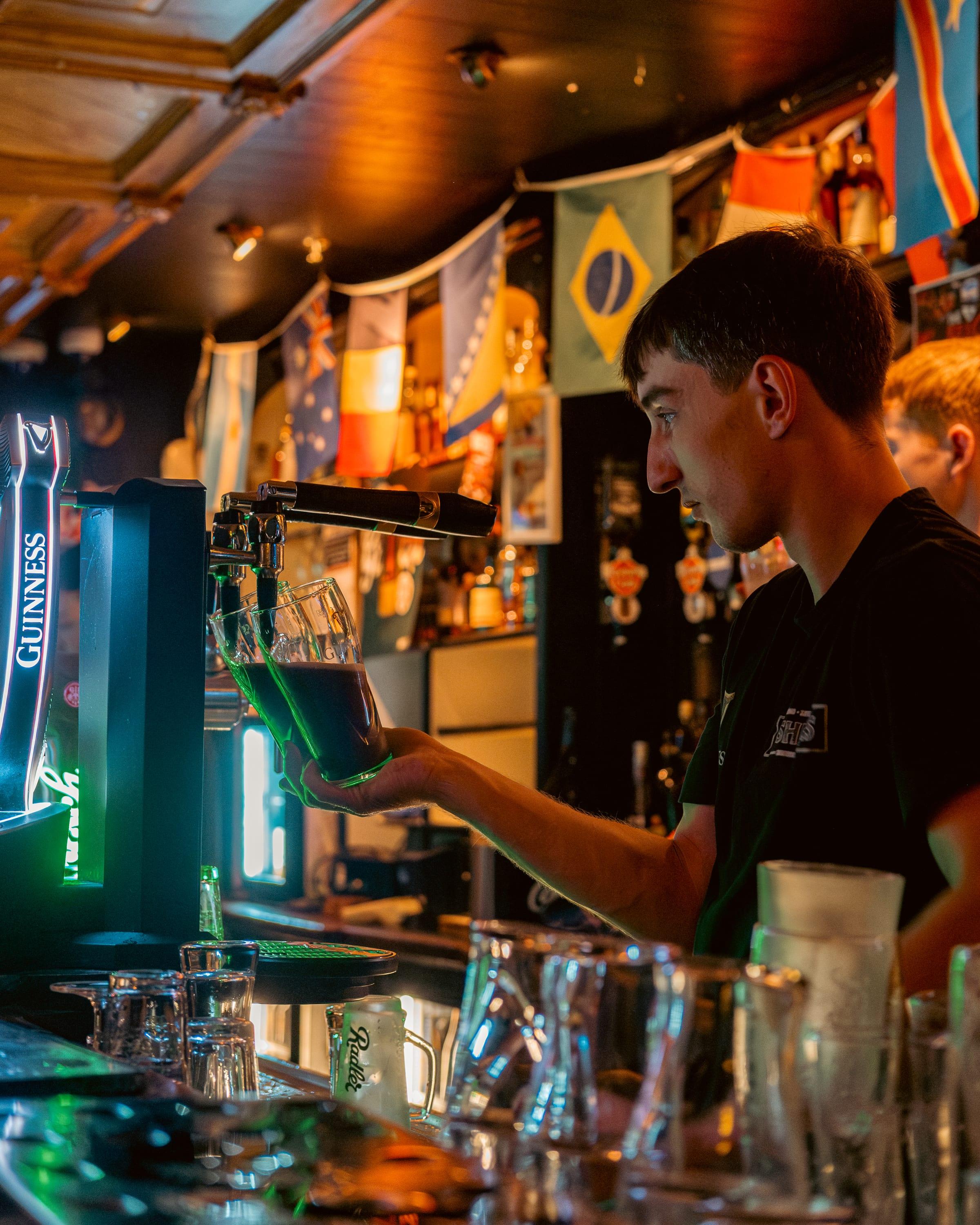 Bartender pulling a pint under green Guinness light, flags strung along the ceiling