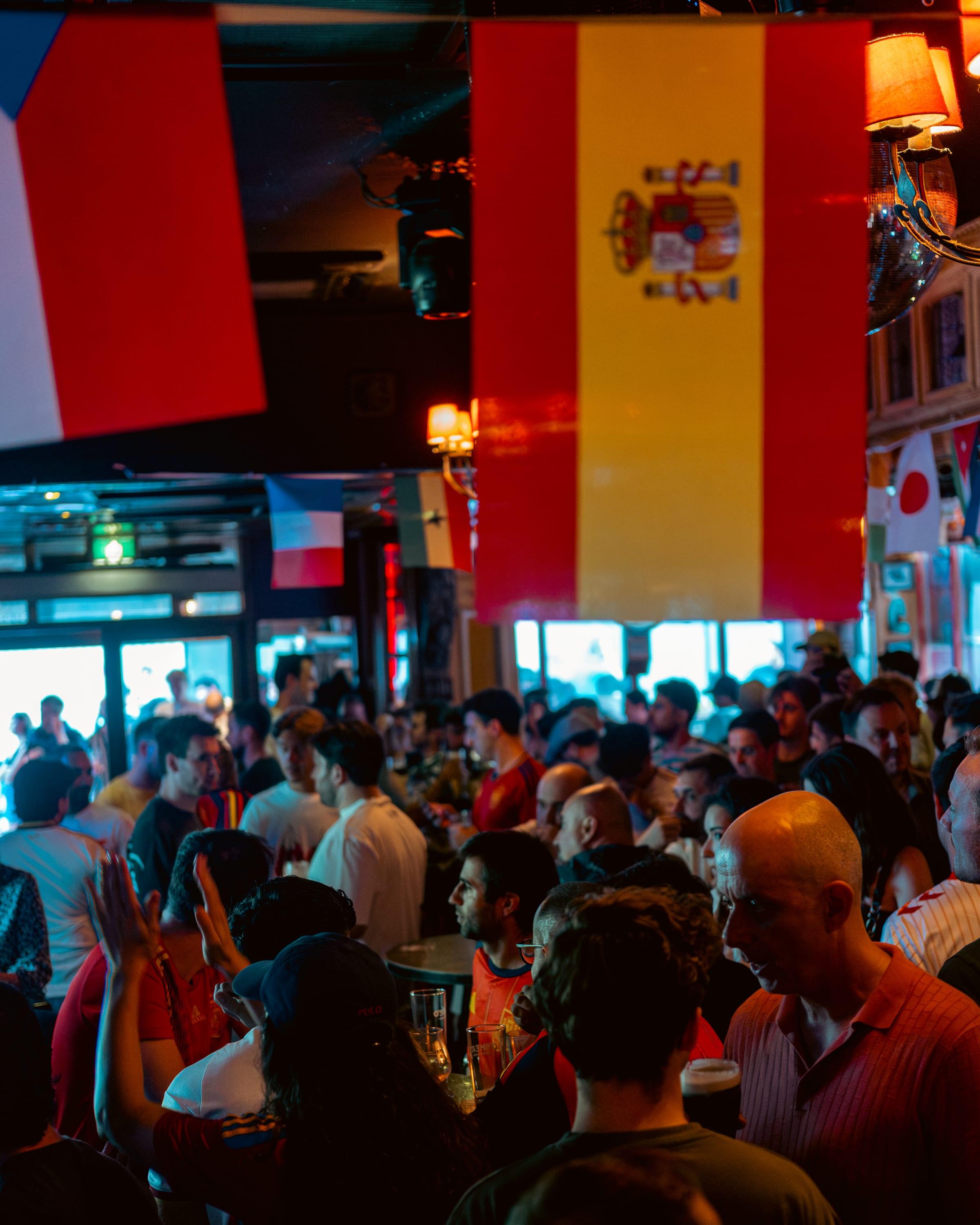 Spain flag hanging low over the pub floor, France flag caught beside it, crowd packed below