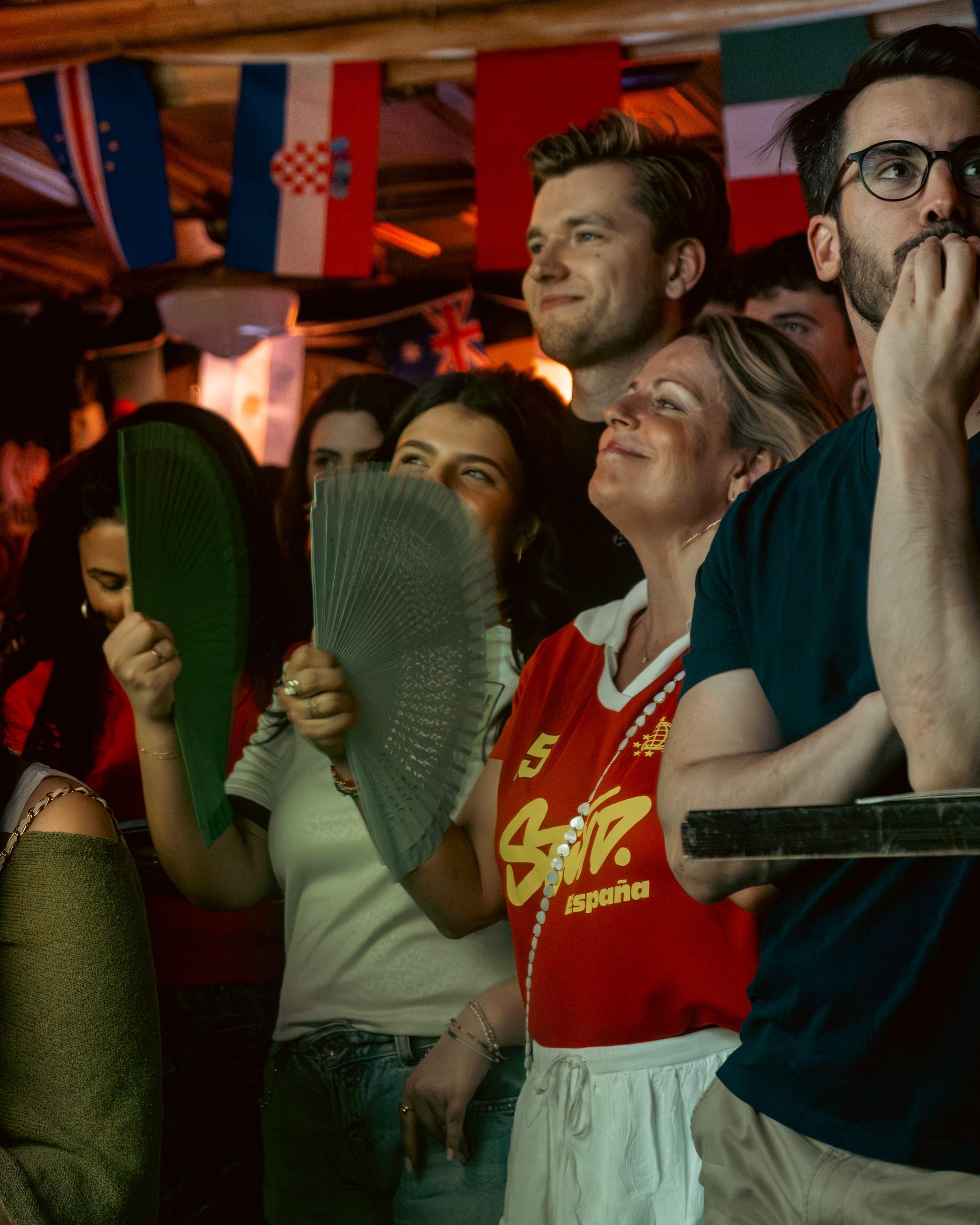 Fans behind paper fans and pint glasses, one in a España shirt grinning up at the screen