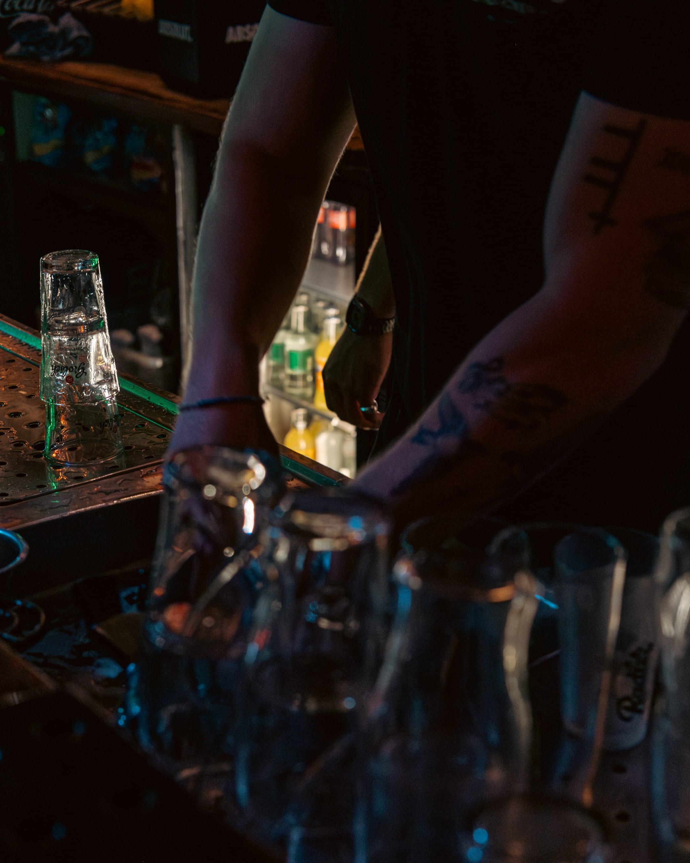 Tattooed forearms and stacked glasses at the bar, caught mid-reach in the rush