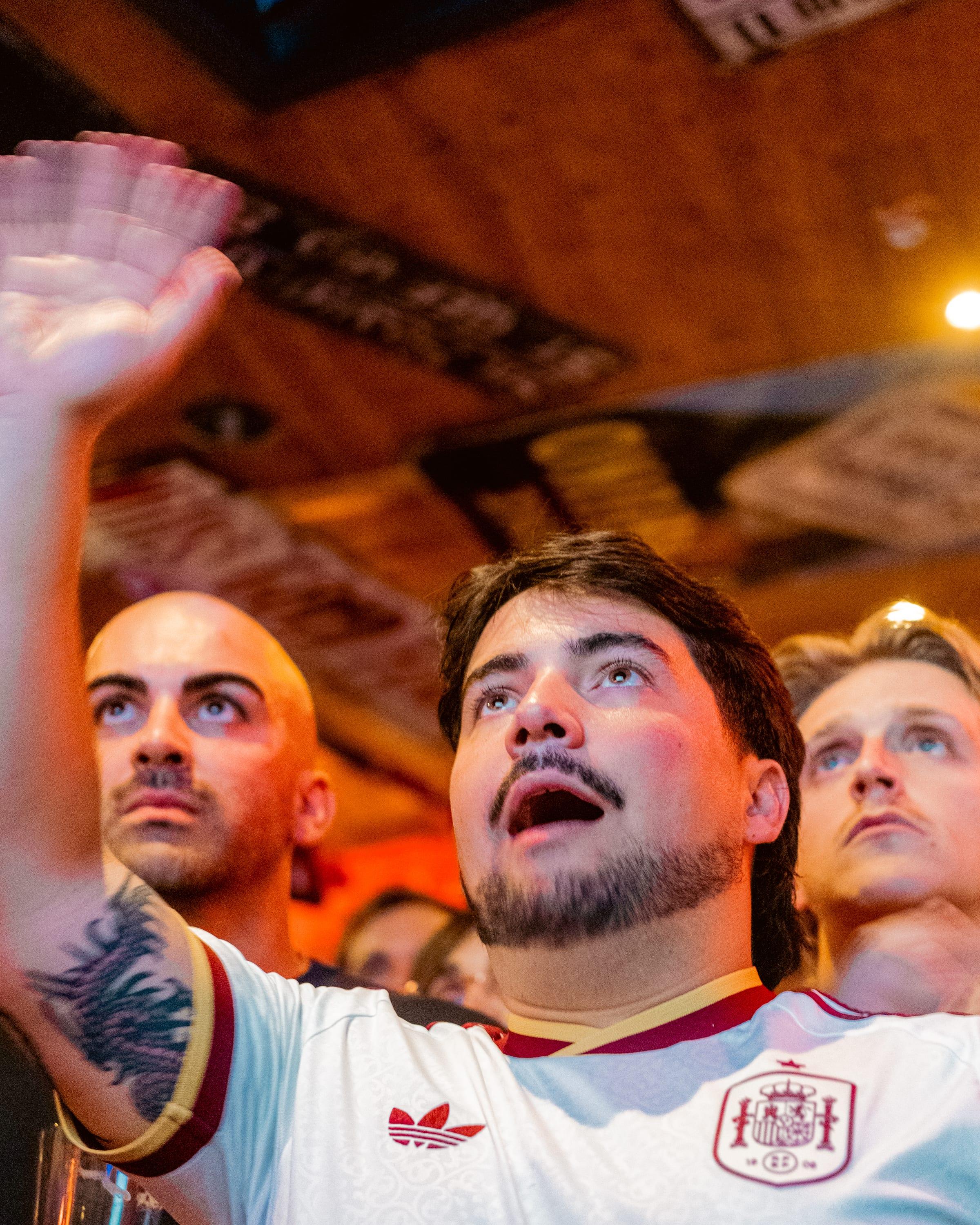 Three fans locked on the screen, one mid-shout in a Spain shirt