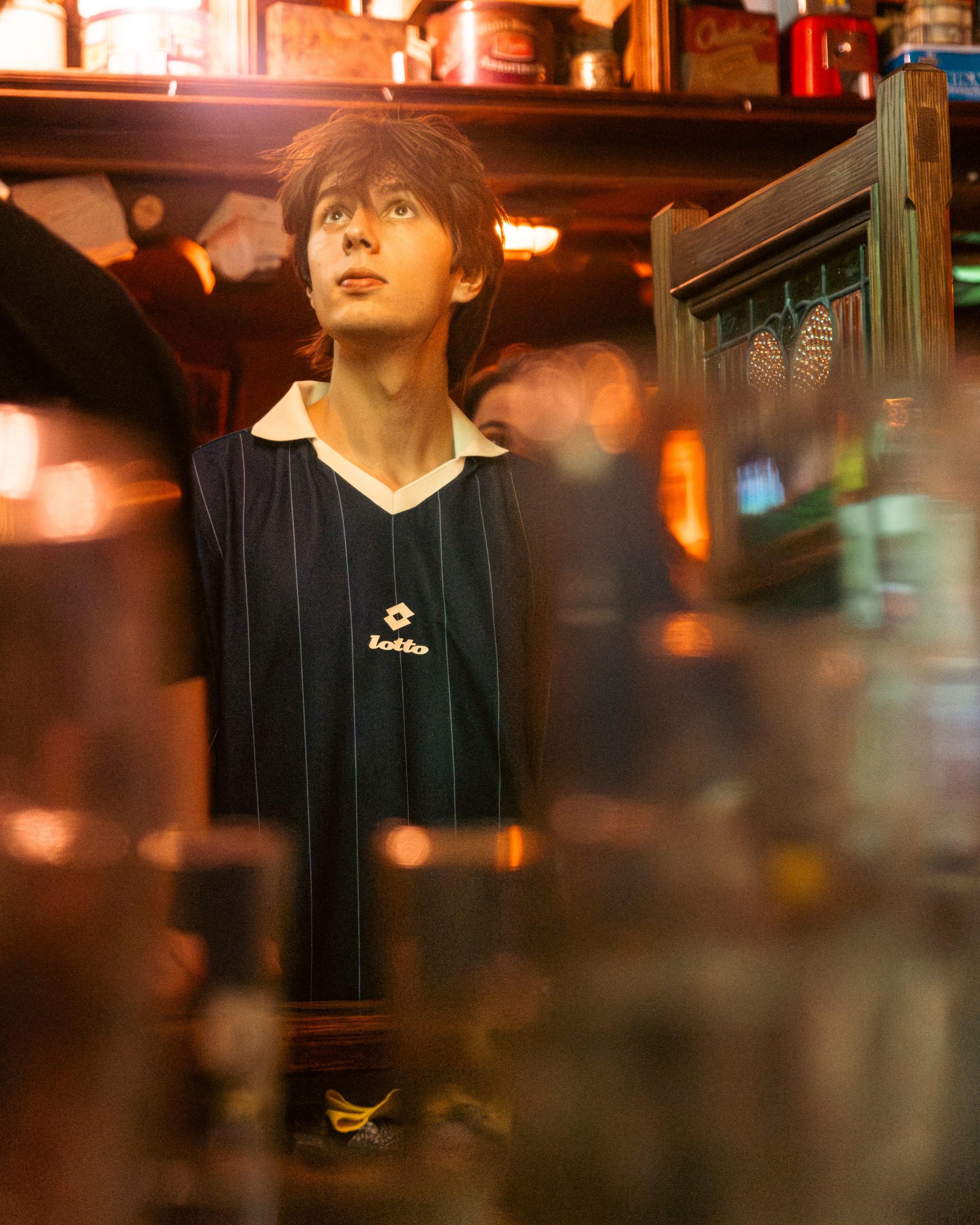 Fan in a vintage navy Lotto shirt looking up at the screen, pub shelves glowing behind him