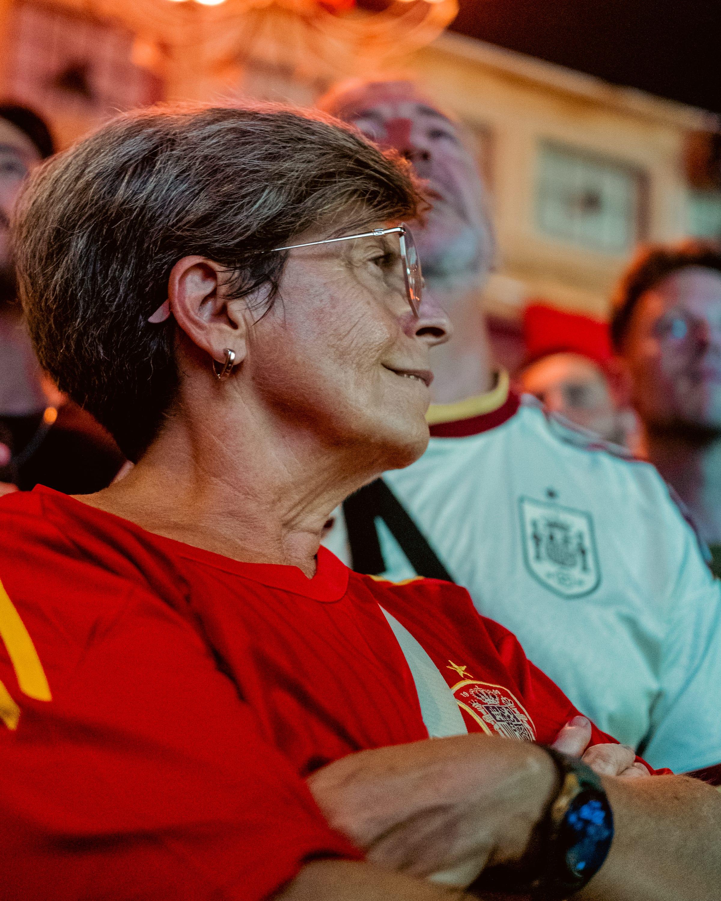 Profile of an older fan in a red Spain shirt, glasses catching the light