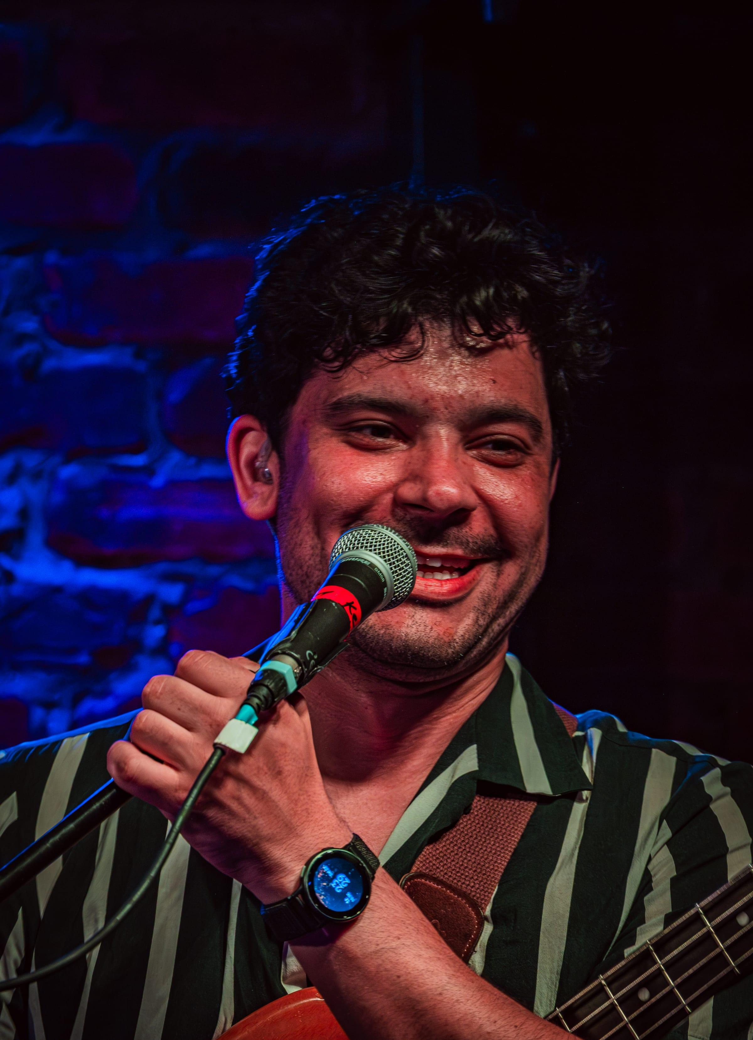 Bassist grinning into the mic between lines, in-ear monitor catching the light, blue brick wall behind him