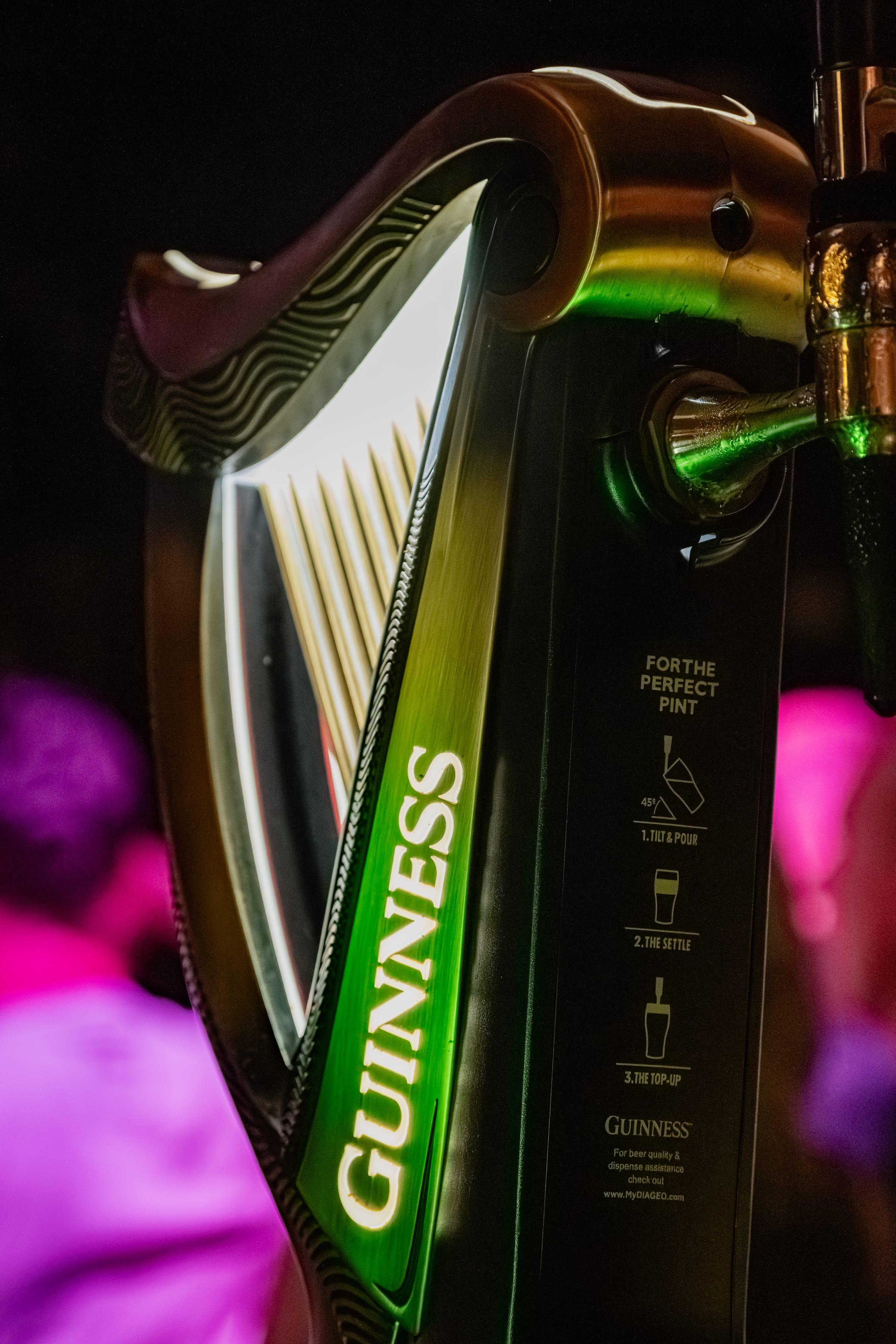 Close detail of a Guinness tap, harp catching light against a blurred crowd in pink and violet