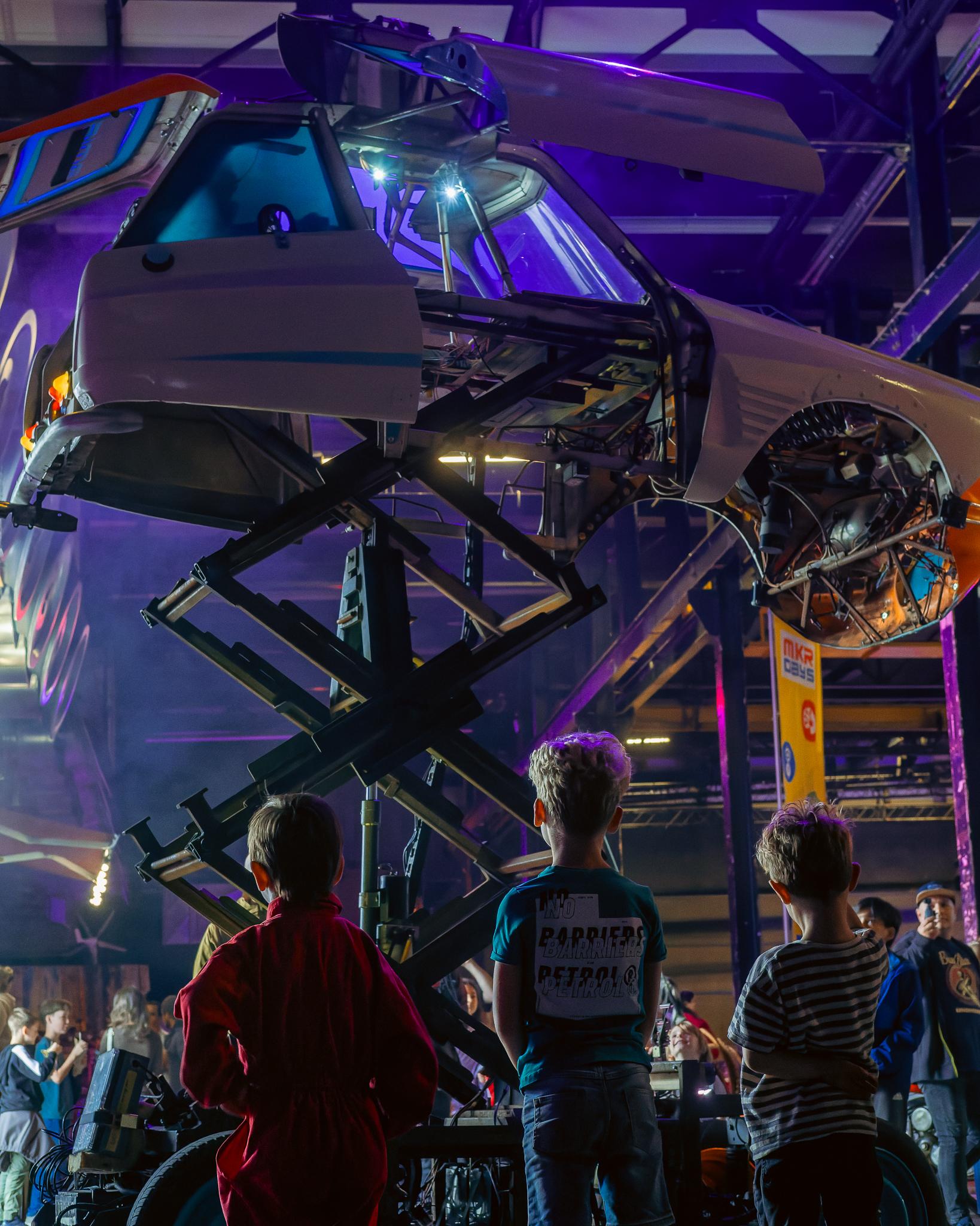 Children looking up at a car suspended overhead, lit in purple, at a Maker Days Eindhoven exhibit