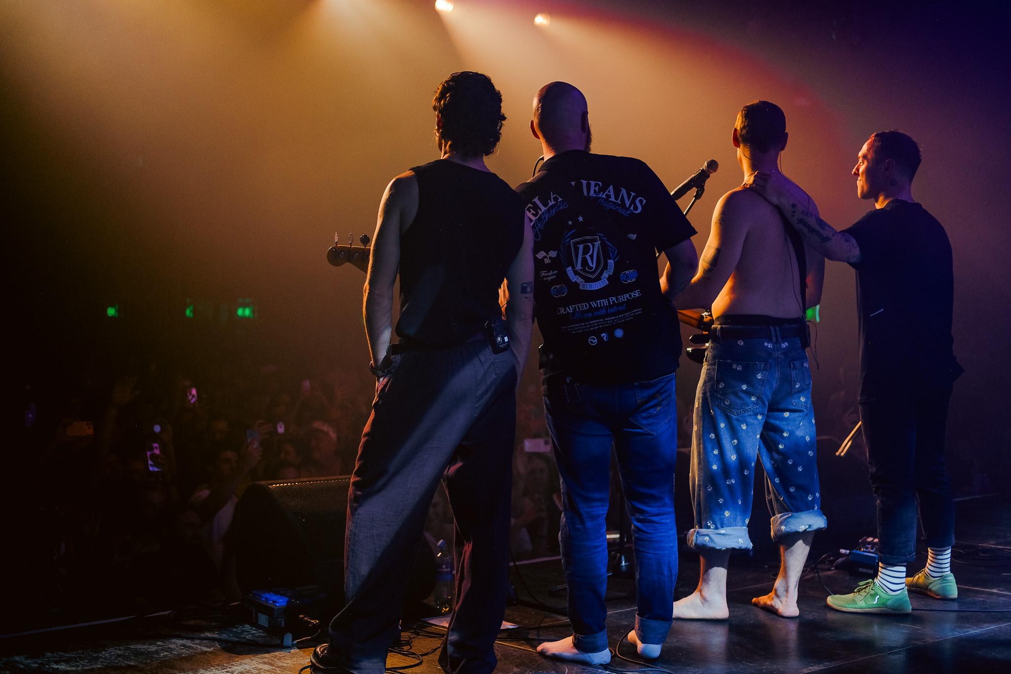 Four band members seen from behind, silhouetted in amber and teal haze, facing the crowd
