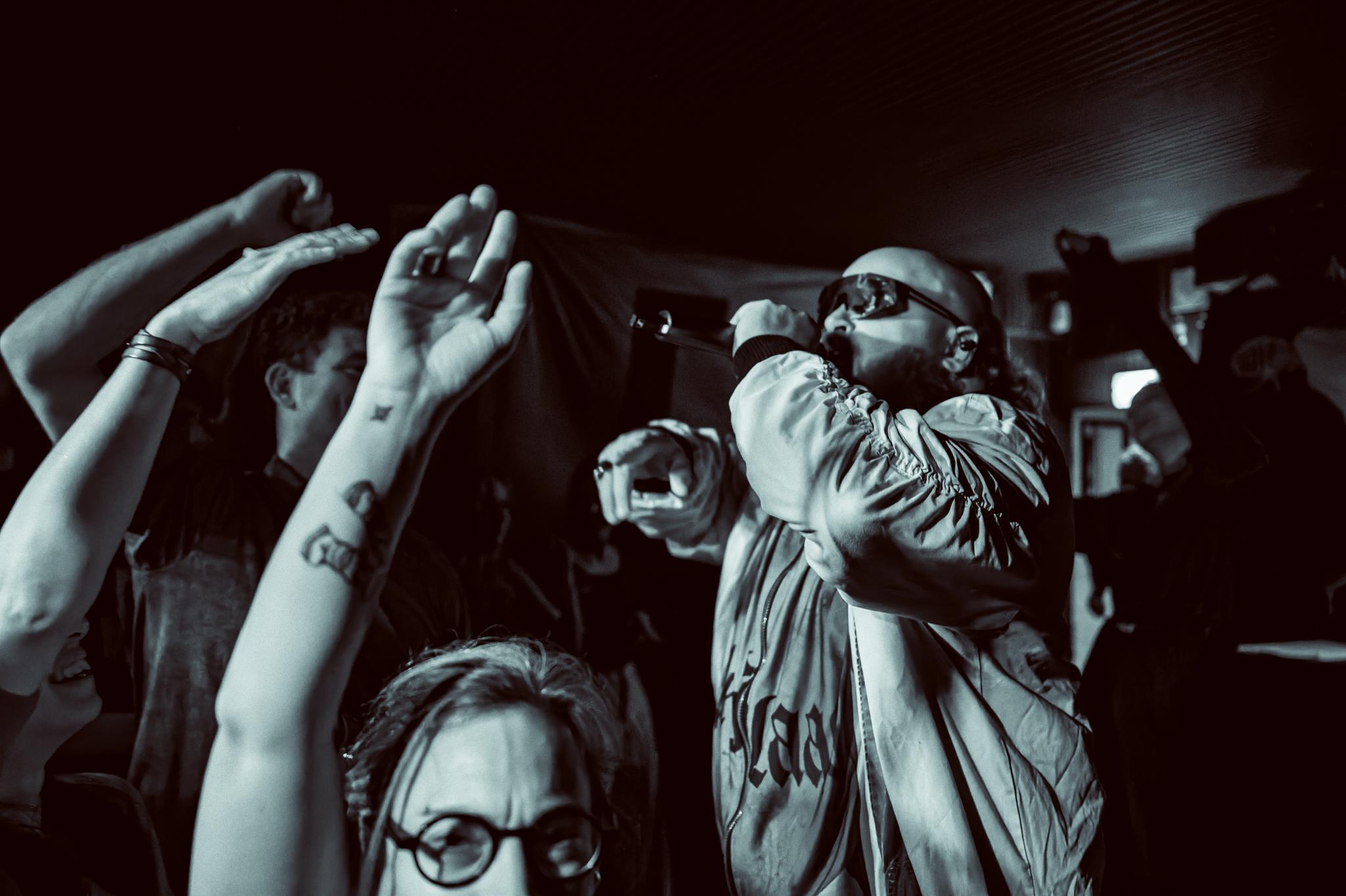 Raised hands in the crowd foreground a singer in sunglasses performing into a mic, black and white