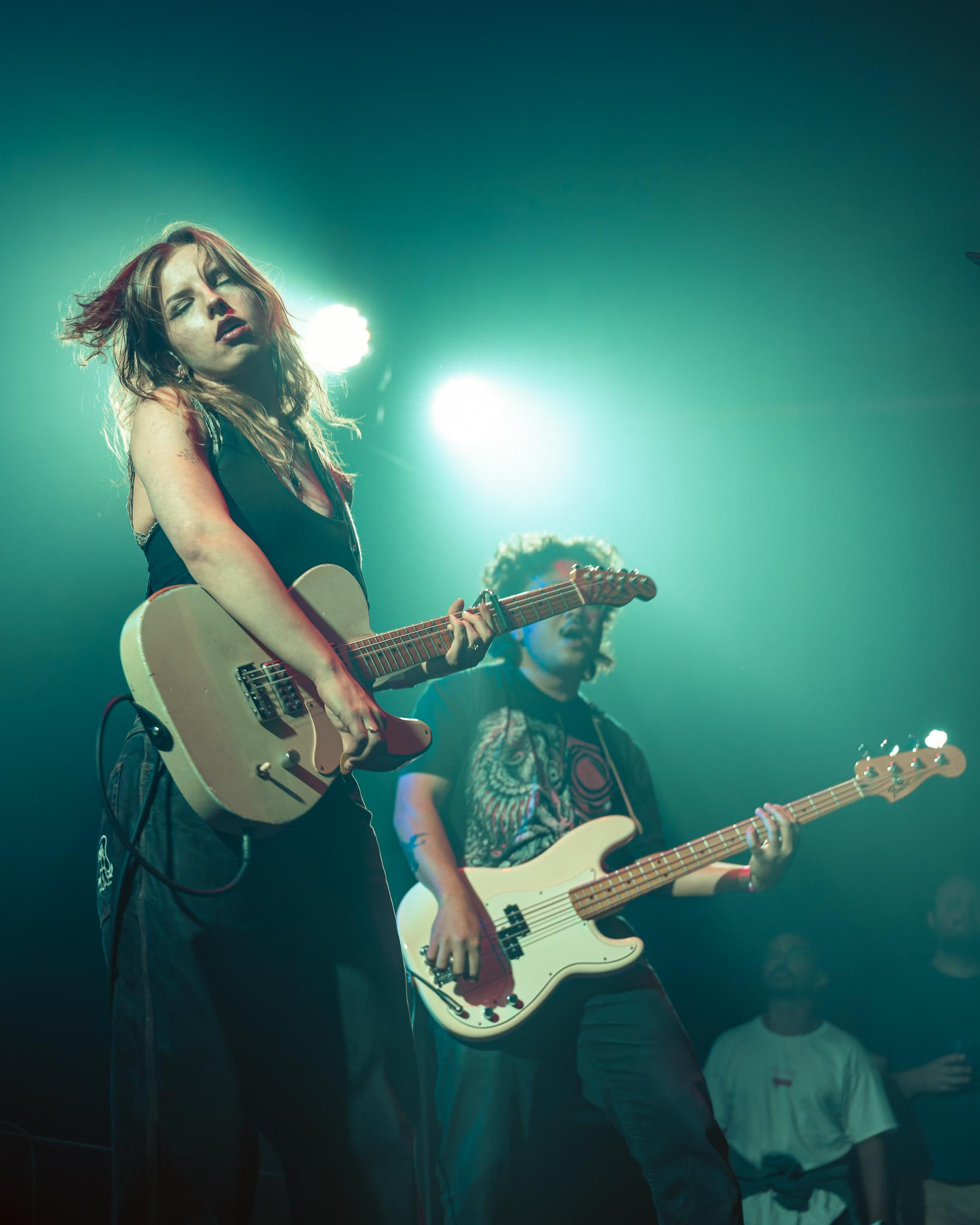A guitarist lost in the moment, eyes closed, bassist playing beside her under teal light