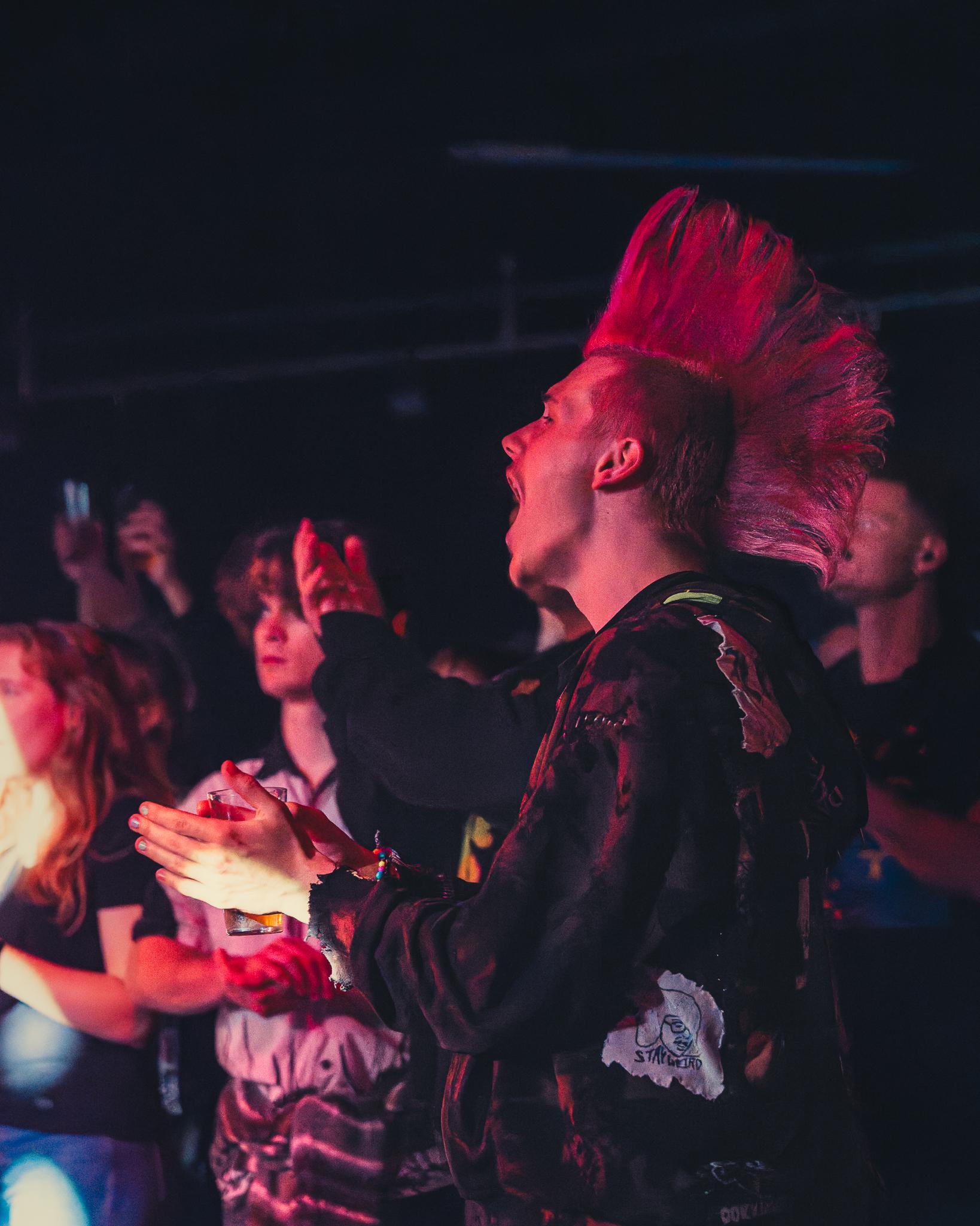 A fan with a pink mohawk cheering mid-crowd, drink raised, under red light