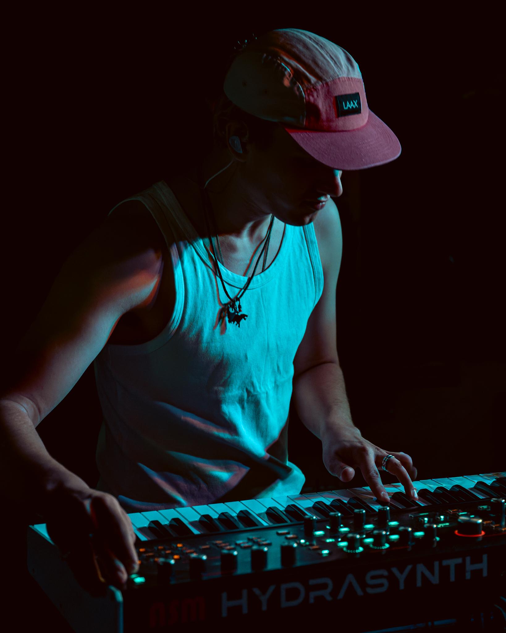 A keyboardist caught in blue light, fingers on the synth, cap pulled low