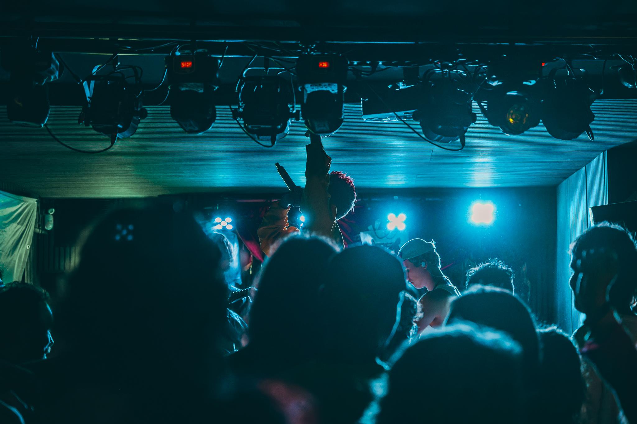 Stage lights rigged overhead, a performer raising a mic above a silhouetted crowd