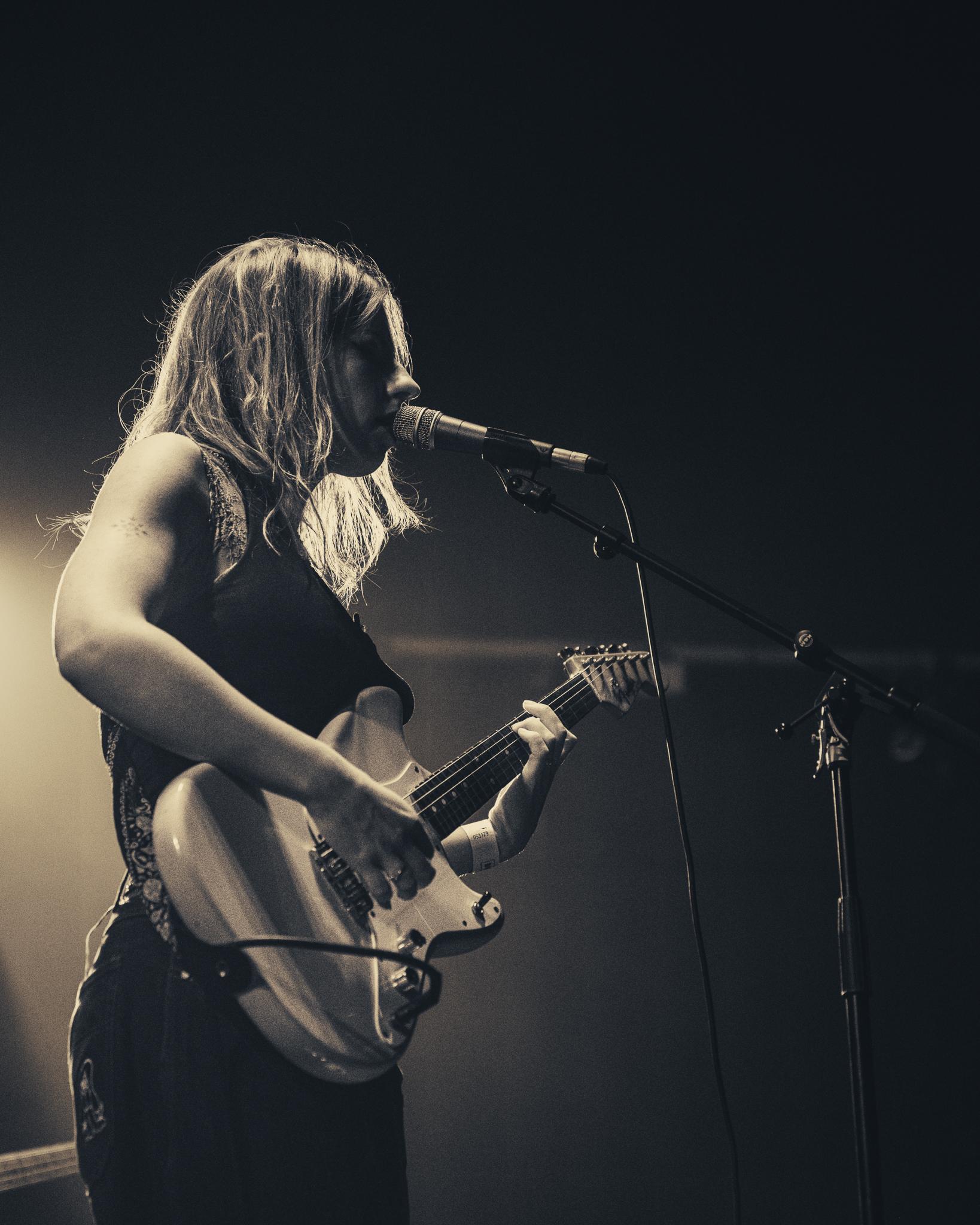 A guitarist singing into the mic, side profile under warm stage light, black and white