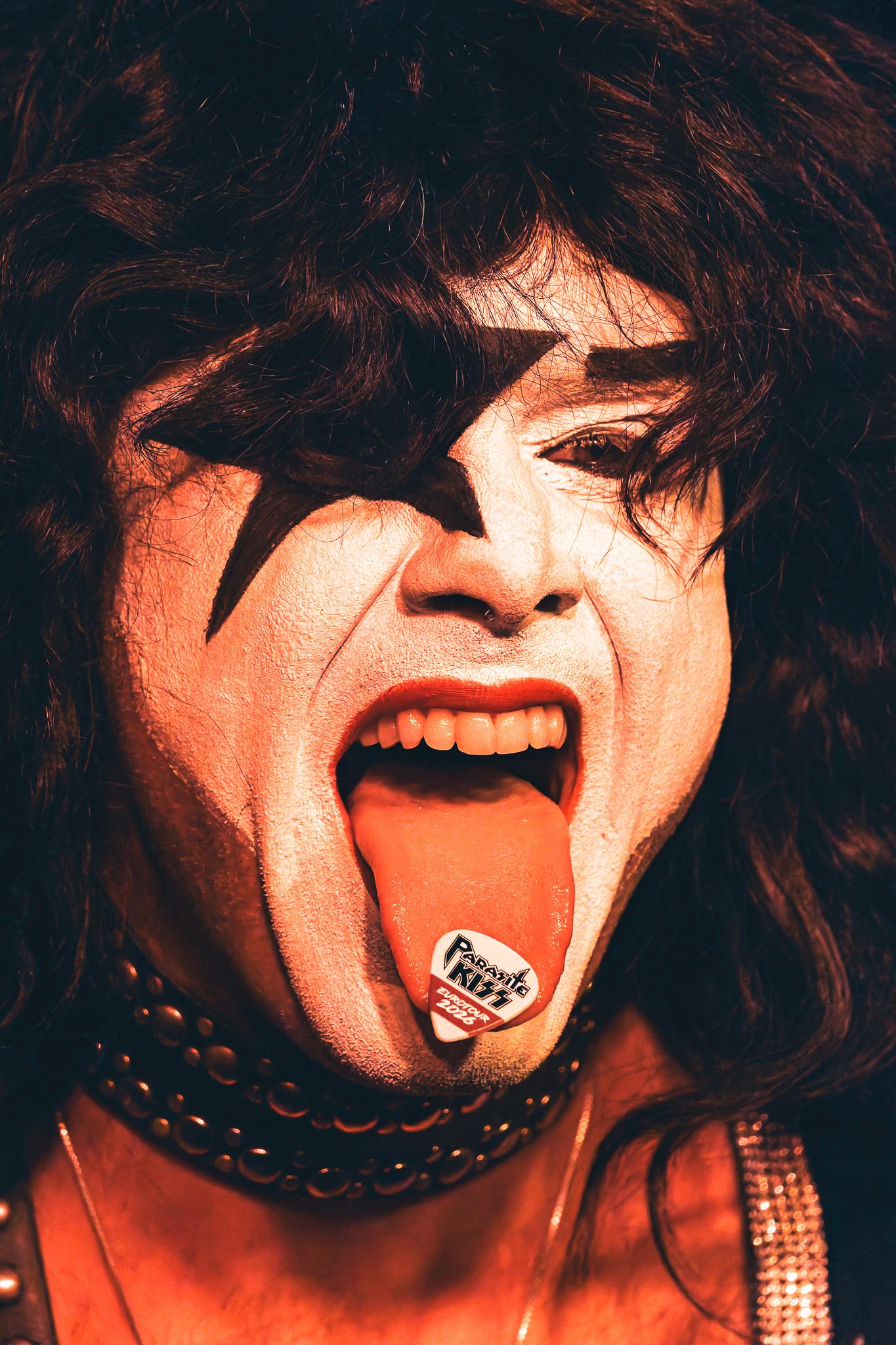 A KISS-style face-painted performer with tongue out, pick balanced on his tongue