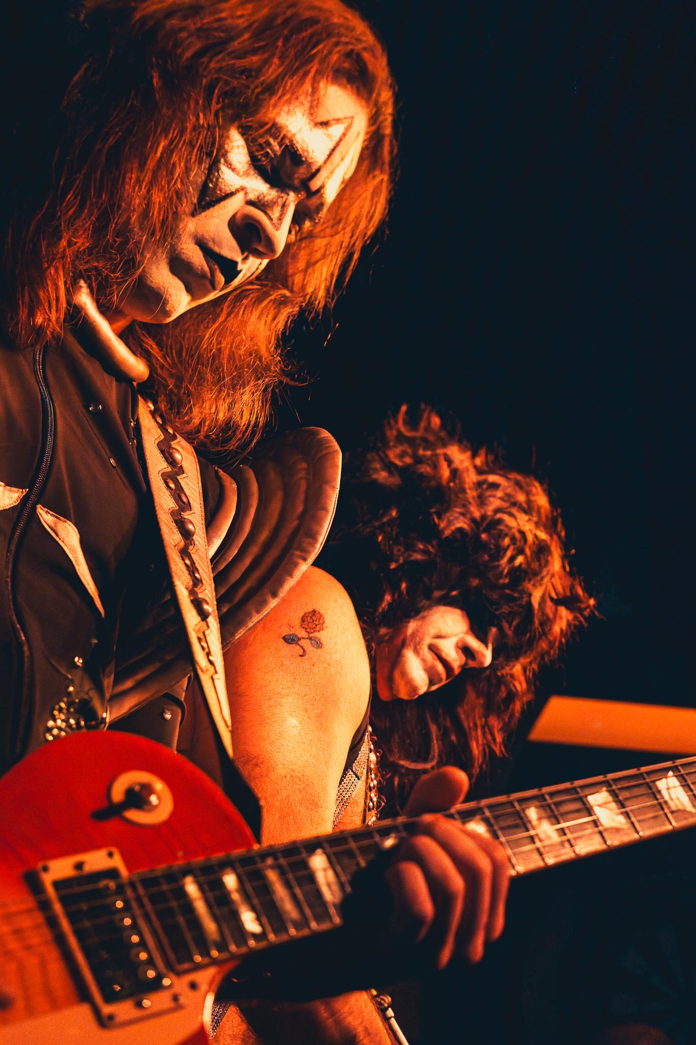 Two KISS-style face-painted guitarists performing, rose tattoo visible on one's leg