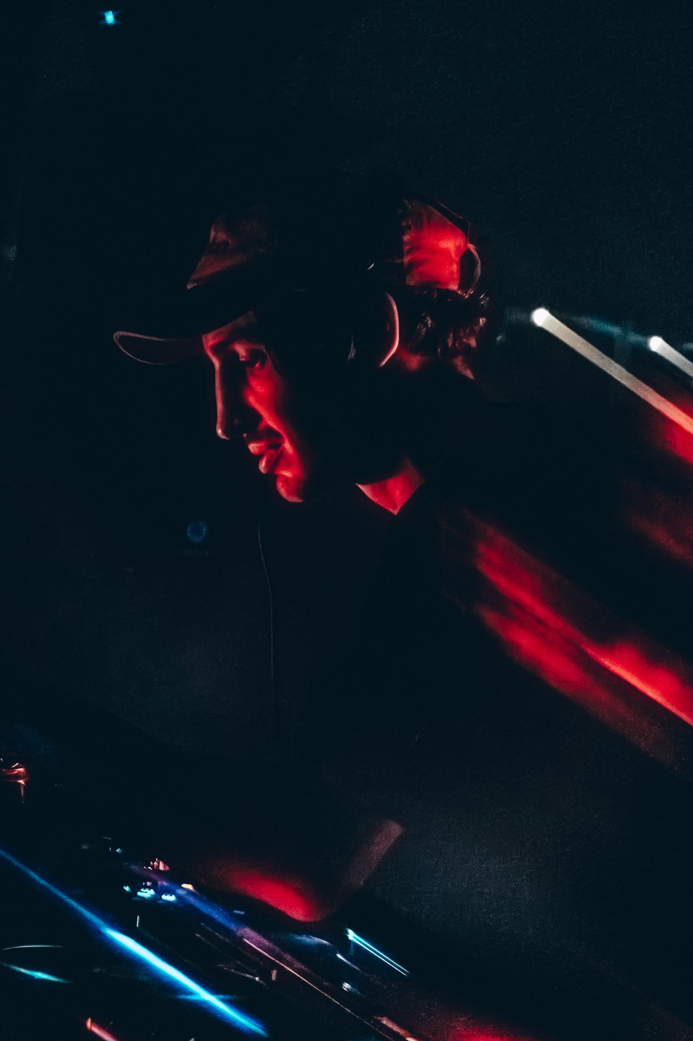A DJ in profile lit in deep red and blue, cap pulled low