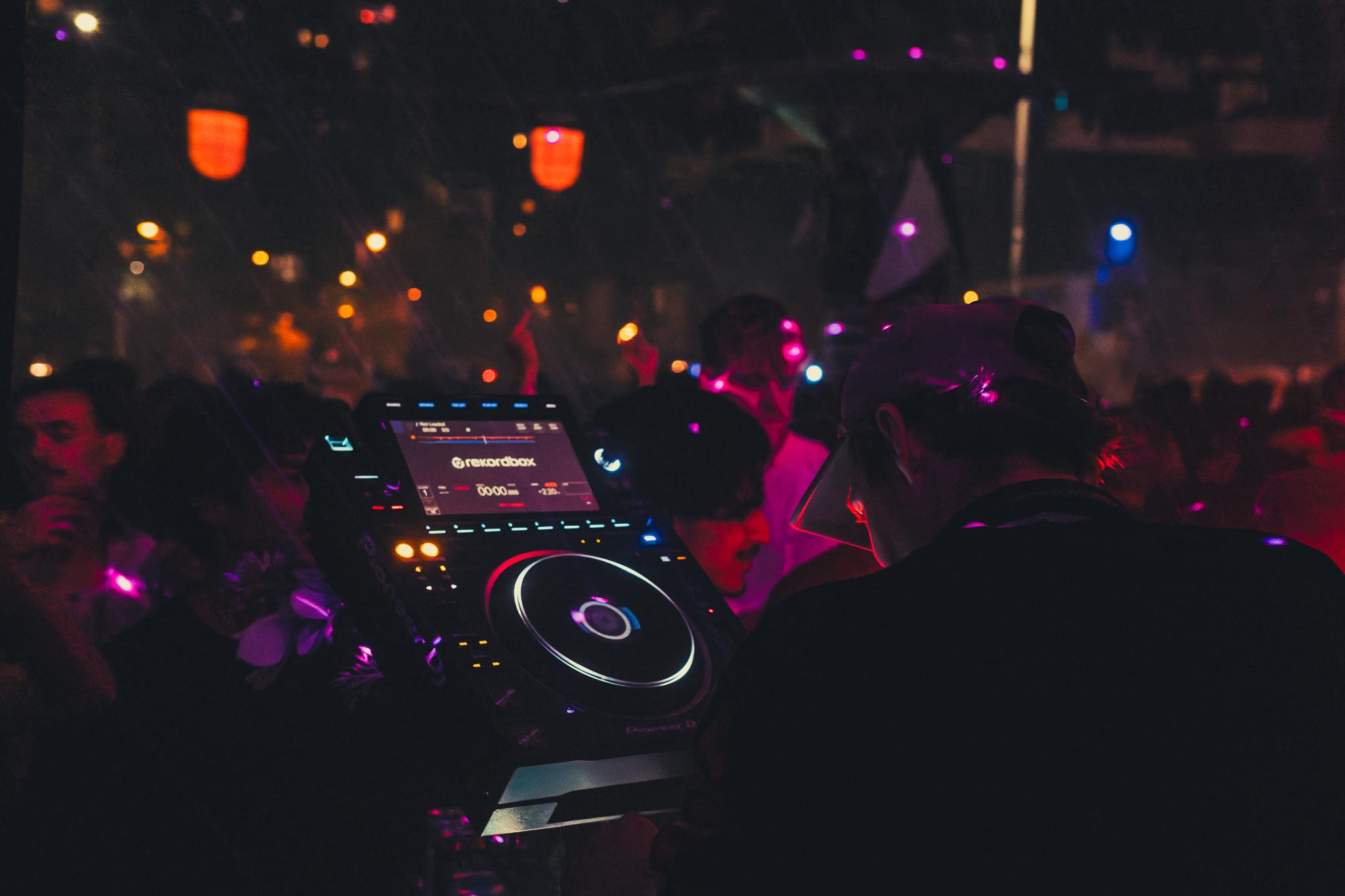 A DJ at the decks, crowd dancing under colourful lights beyond