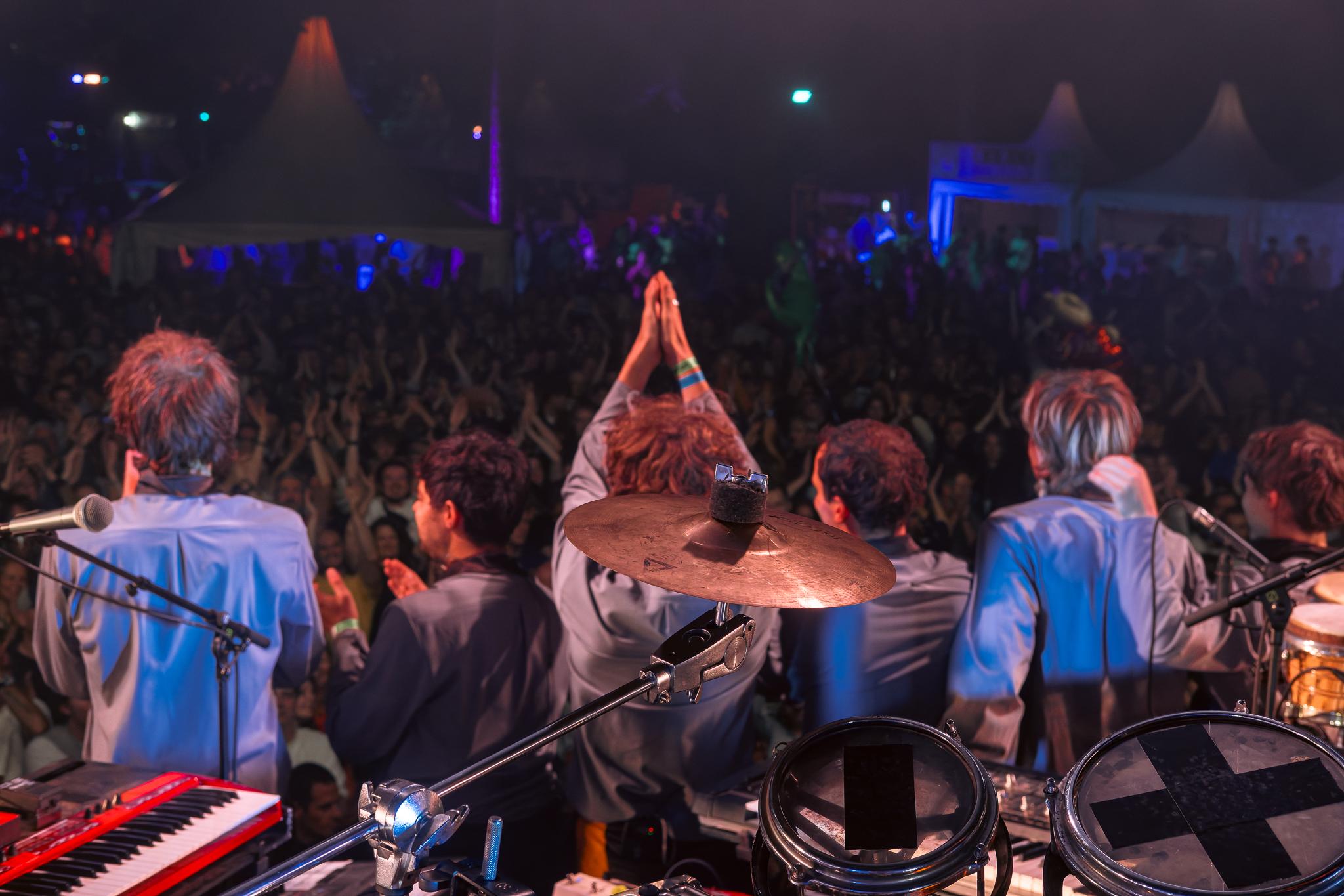 A band seen from behind, arms raised, facing a packed crowd under festival tents