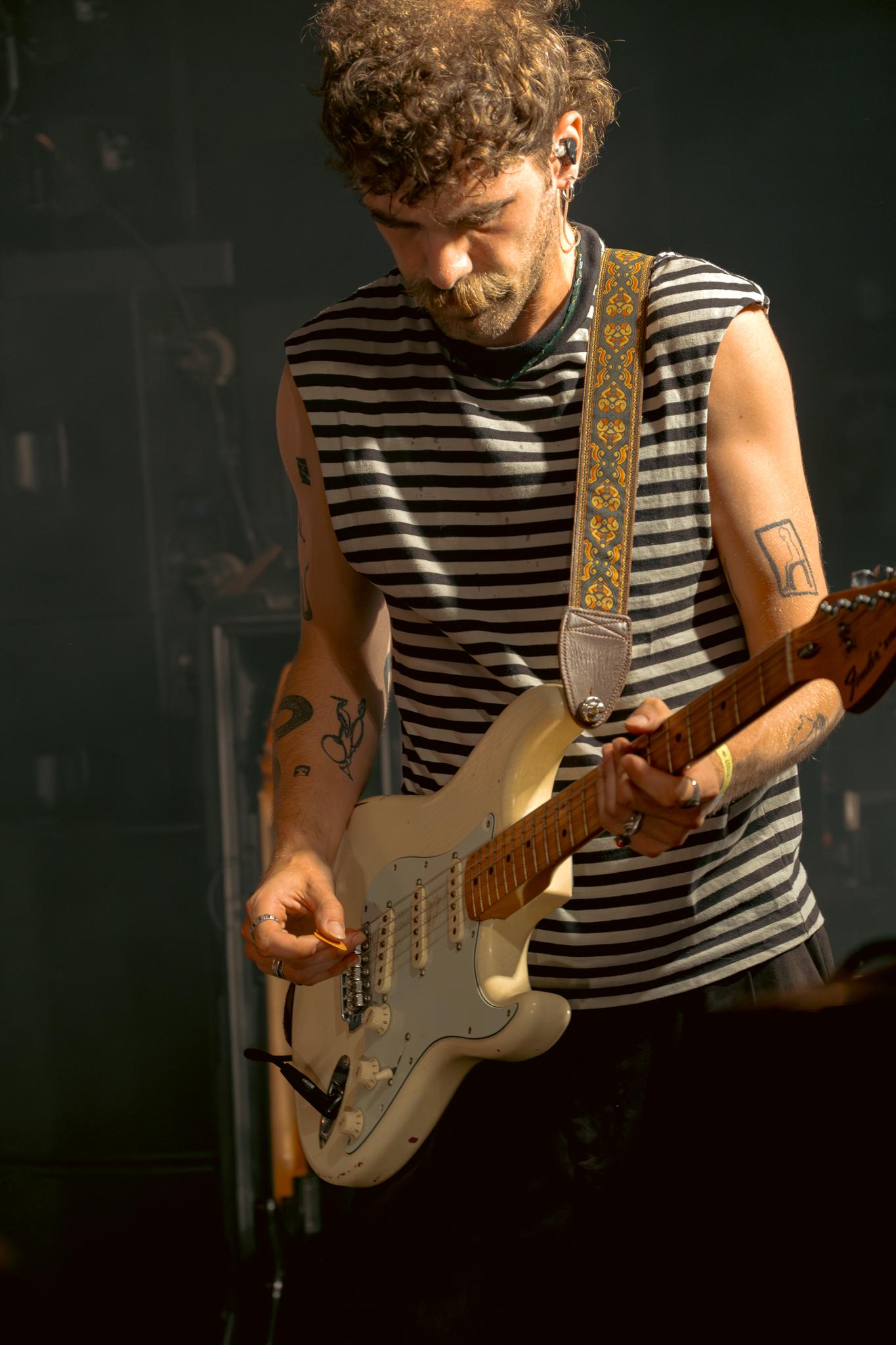 A guitarist in a striped shirt, head down, focused on his white Stratocaster