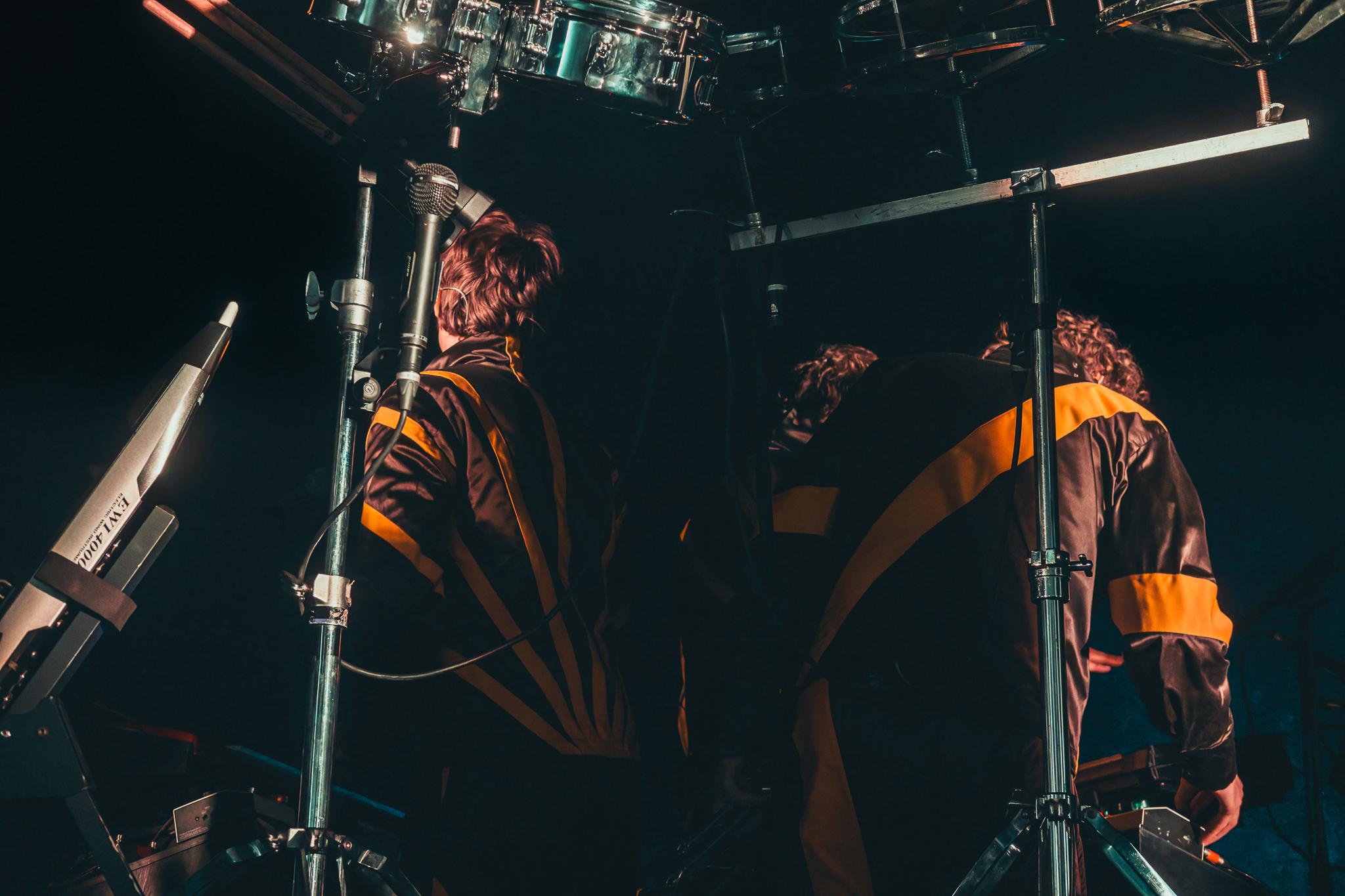 A band viewed from behind under the drum rig, facing a packed festival crowd
