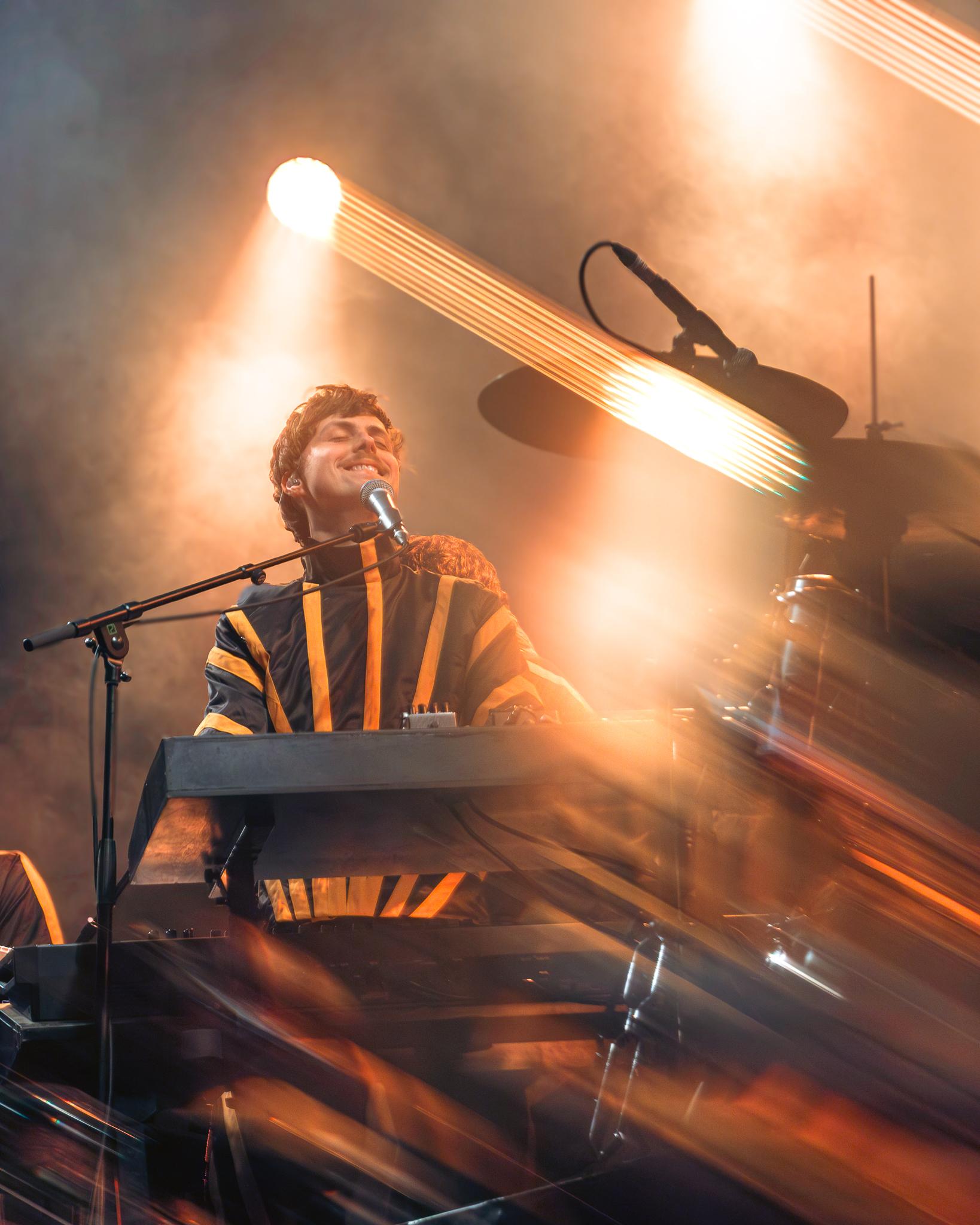 A keyboardist smiling with eyes closed, warm light streaking across the frame