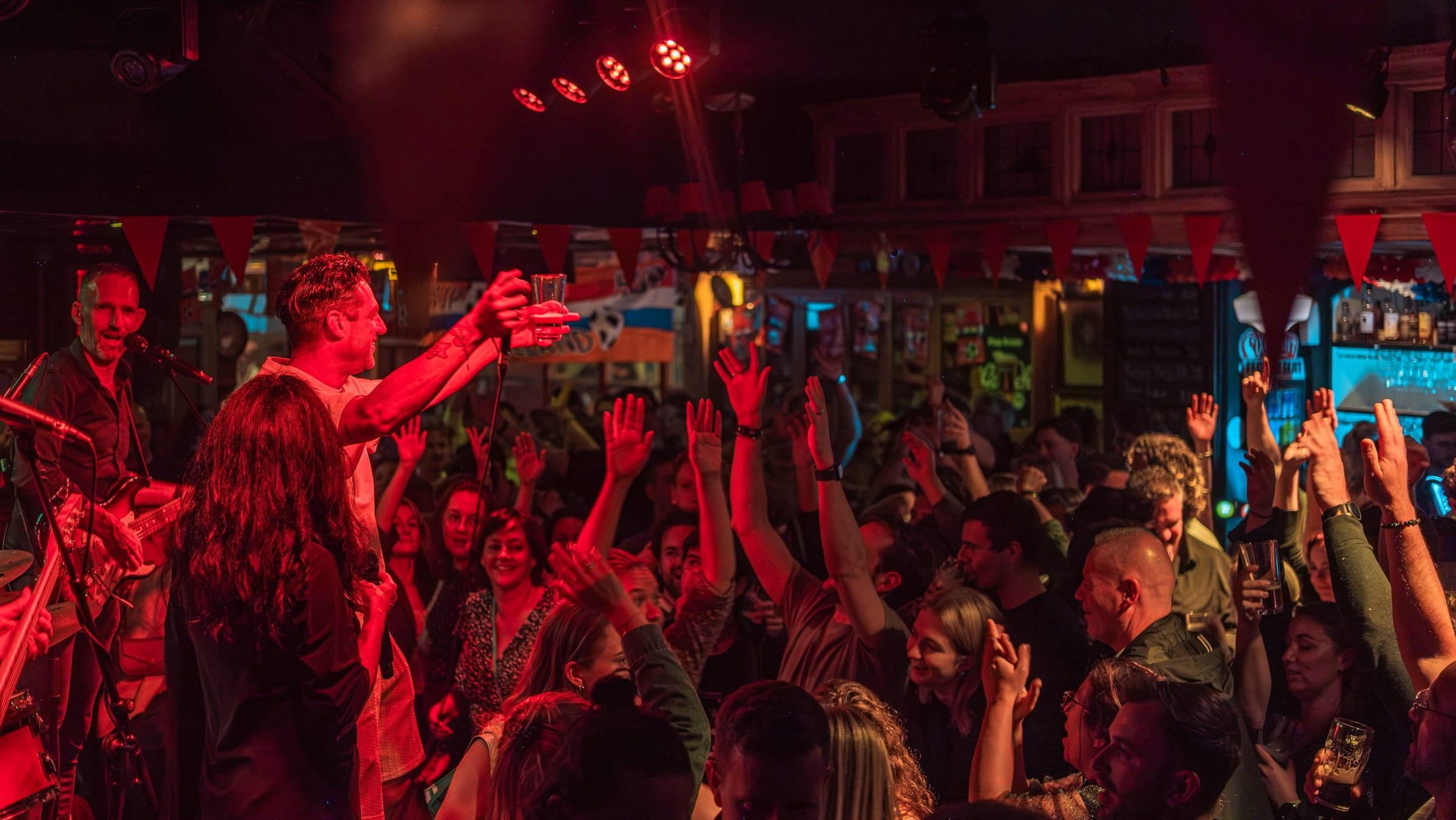 Singer raised on the crowd's shoulders, glass held high, a sea of arms raised beneath red stage light and bunting