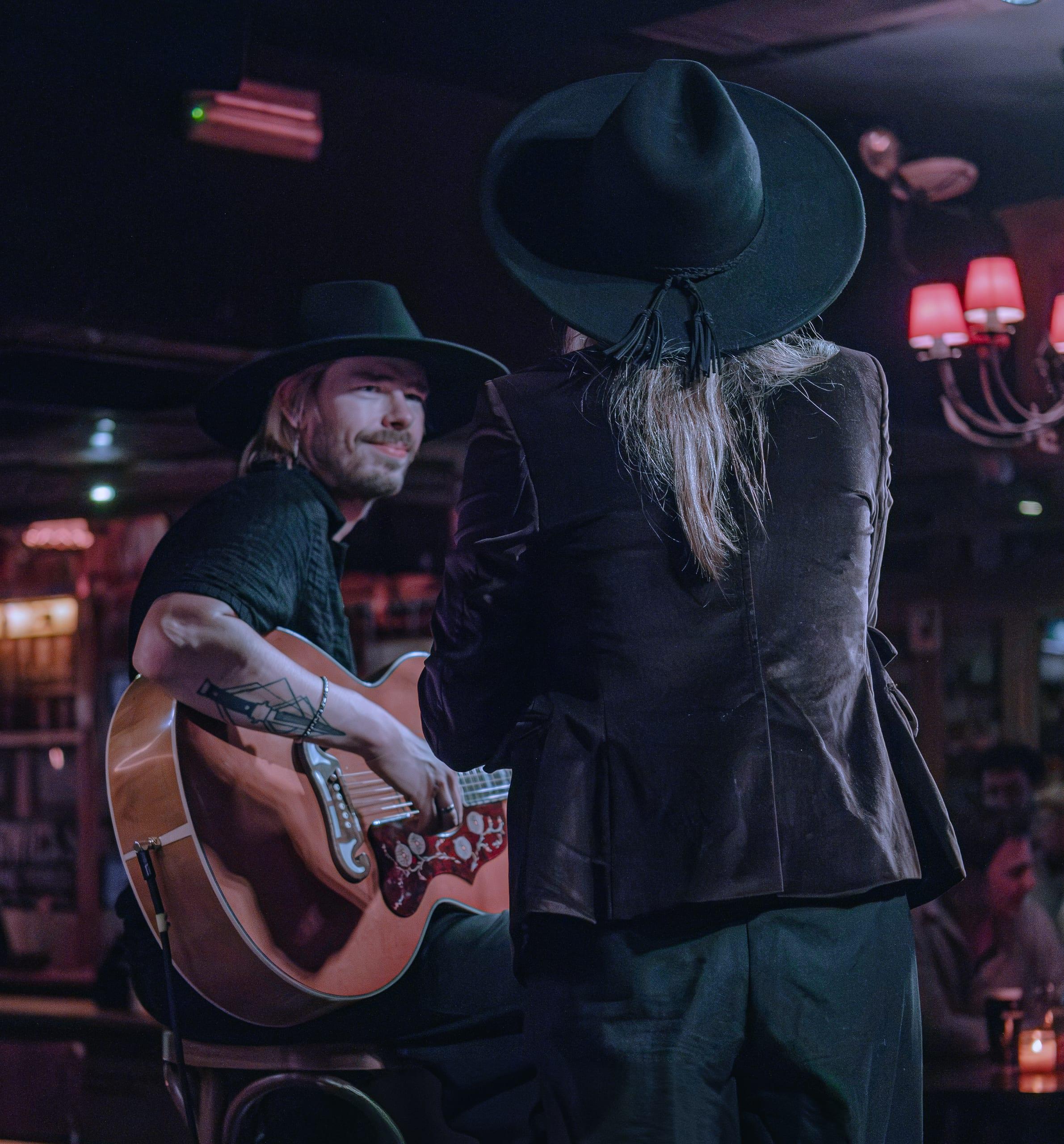 Two performers trading a look mid-song, one on acoustic guitar, under a chandelier's red light