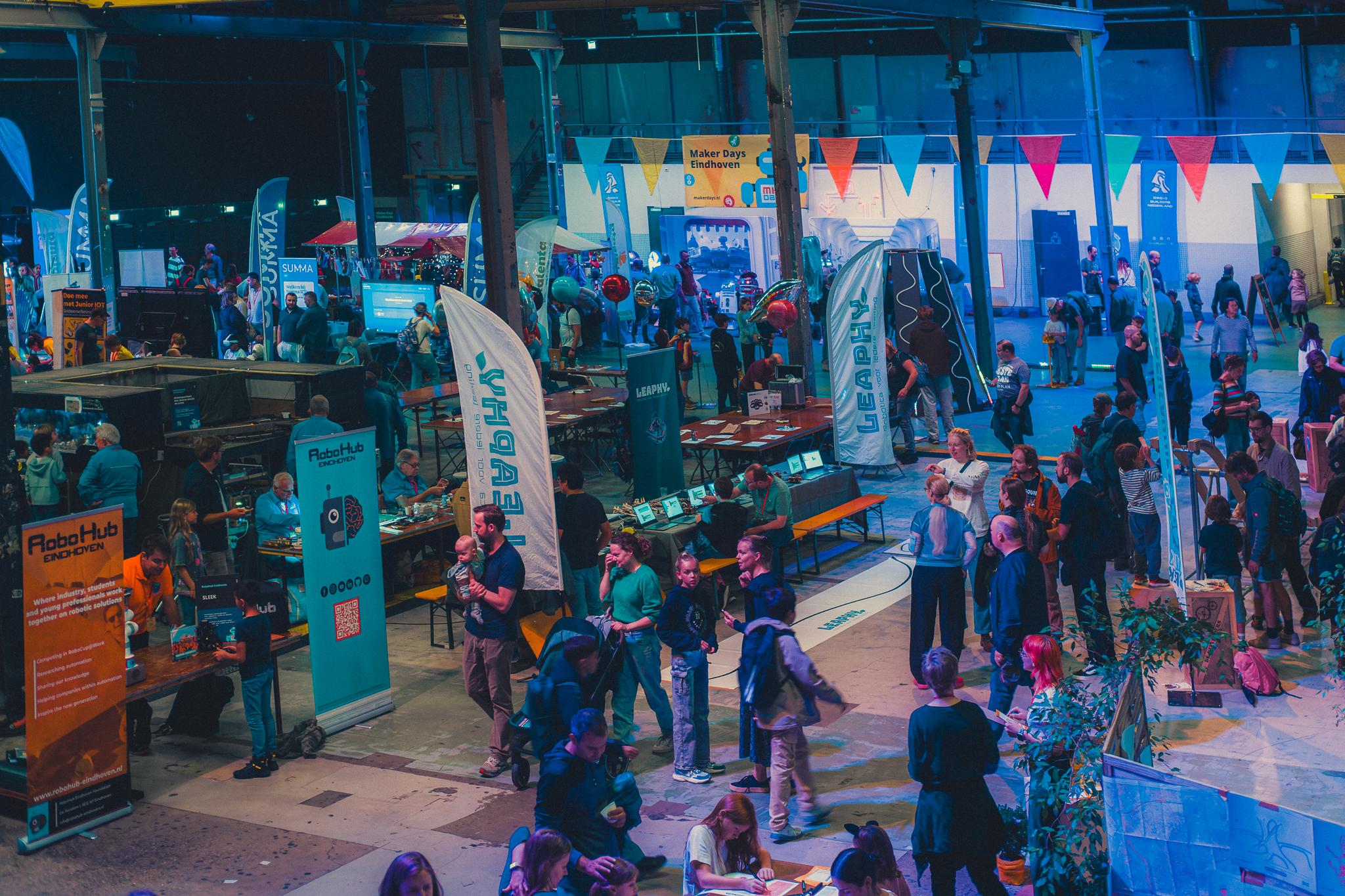 Wide view of the Maker Days Eindhoven exhibition hall, banners and stands under blue light
