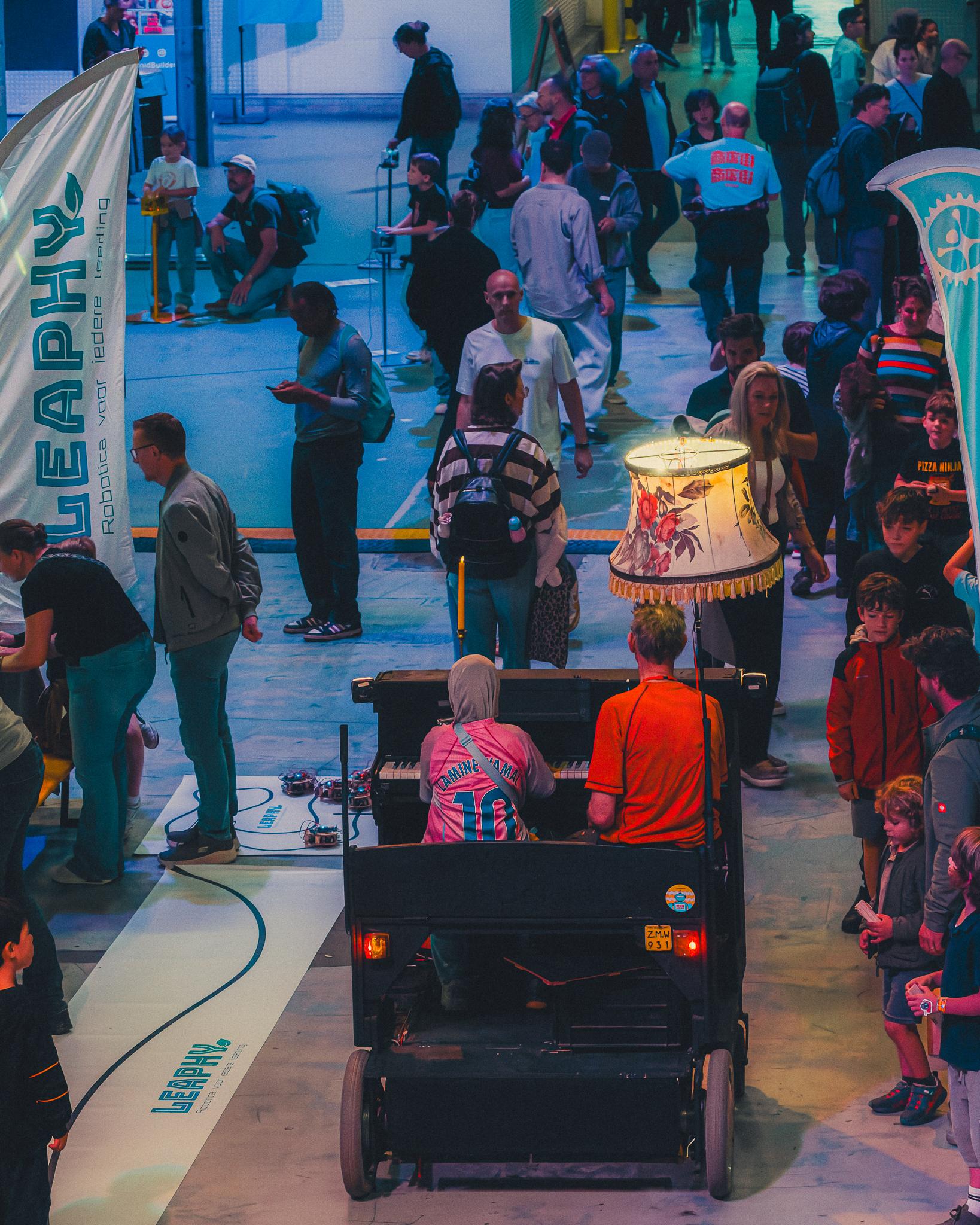 Buskers playing a wheeled piano cart while a crowd browses robotics stands behind them