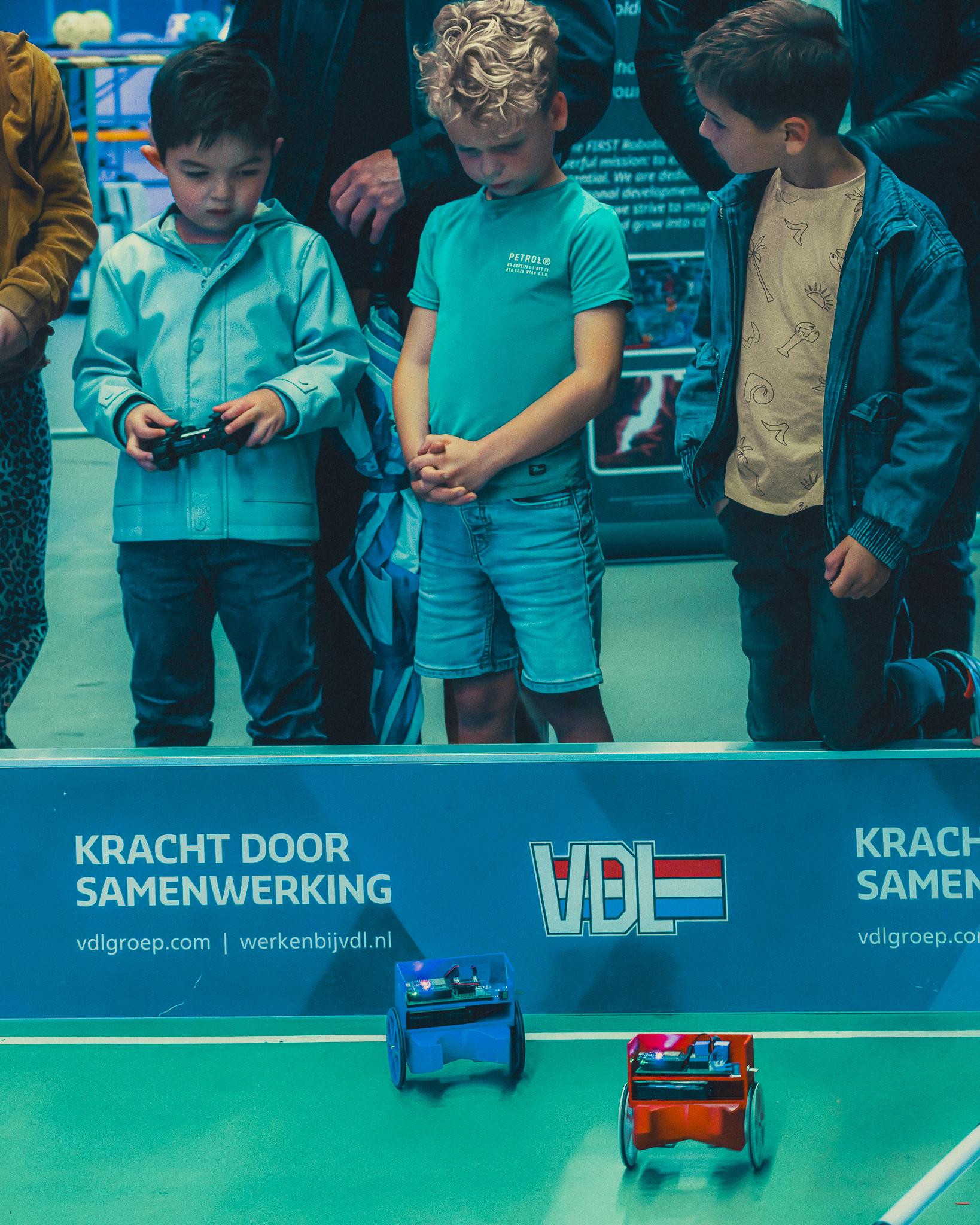 Three boys watching two small remote-control robot cars face off in front of a VDL banner