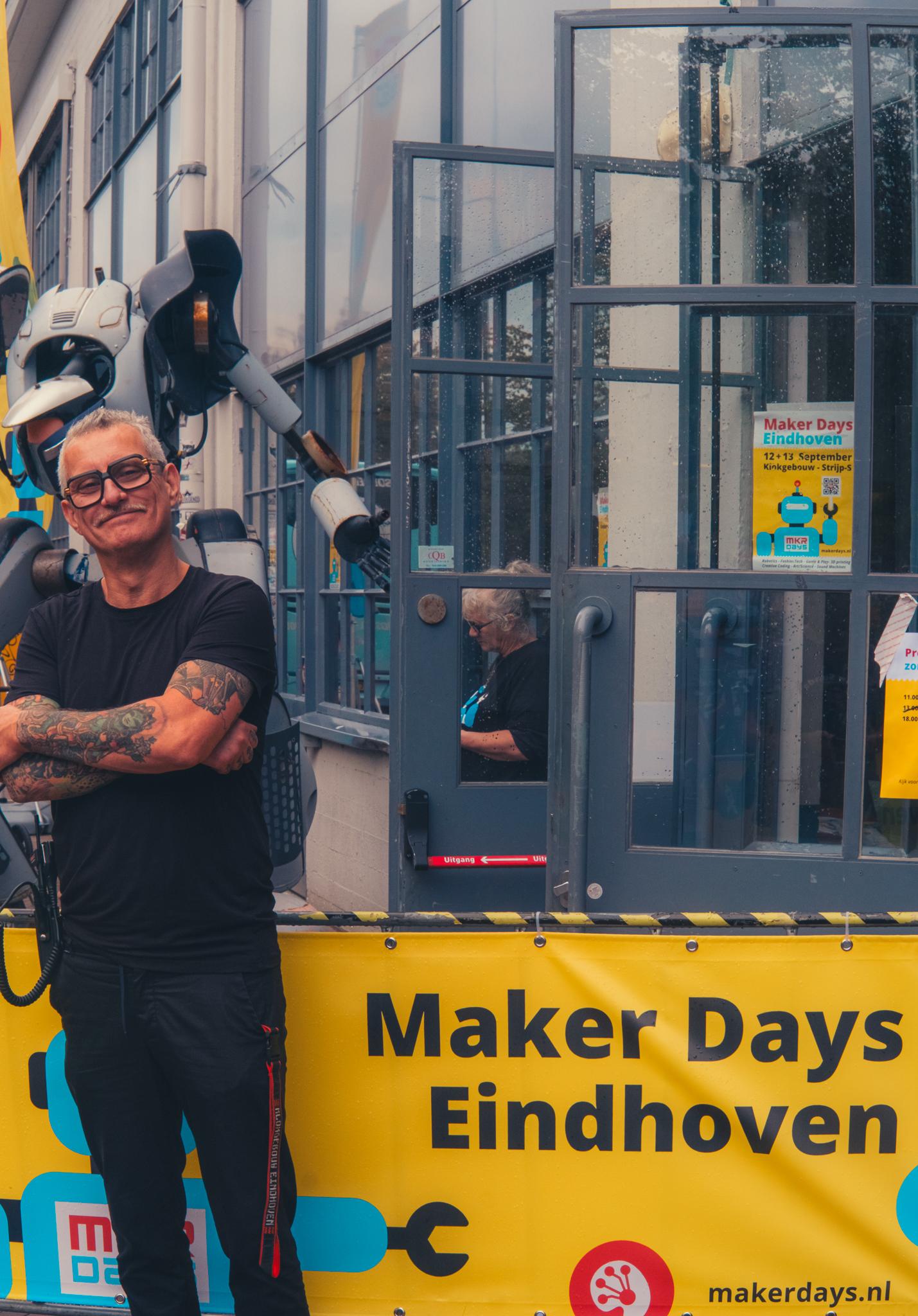 Tattooed RoboHub staff member smiling in front of the Maker Days Eindhoven banner