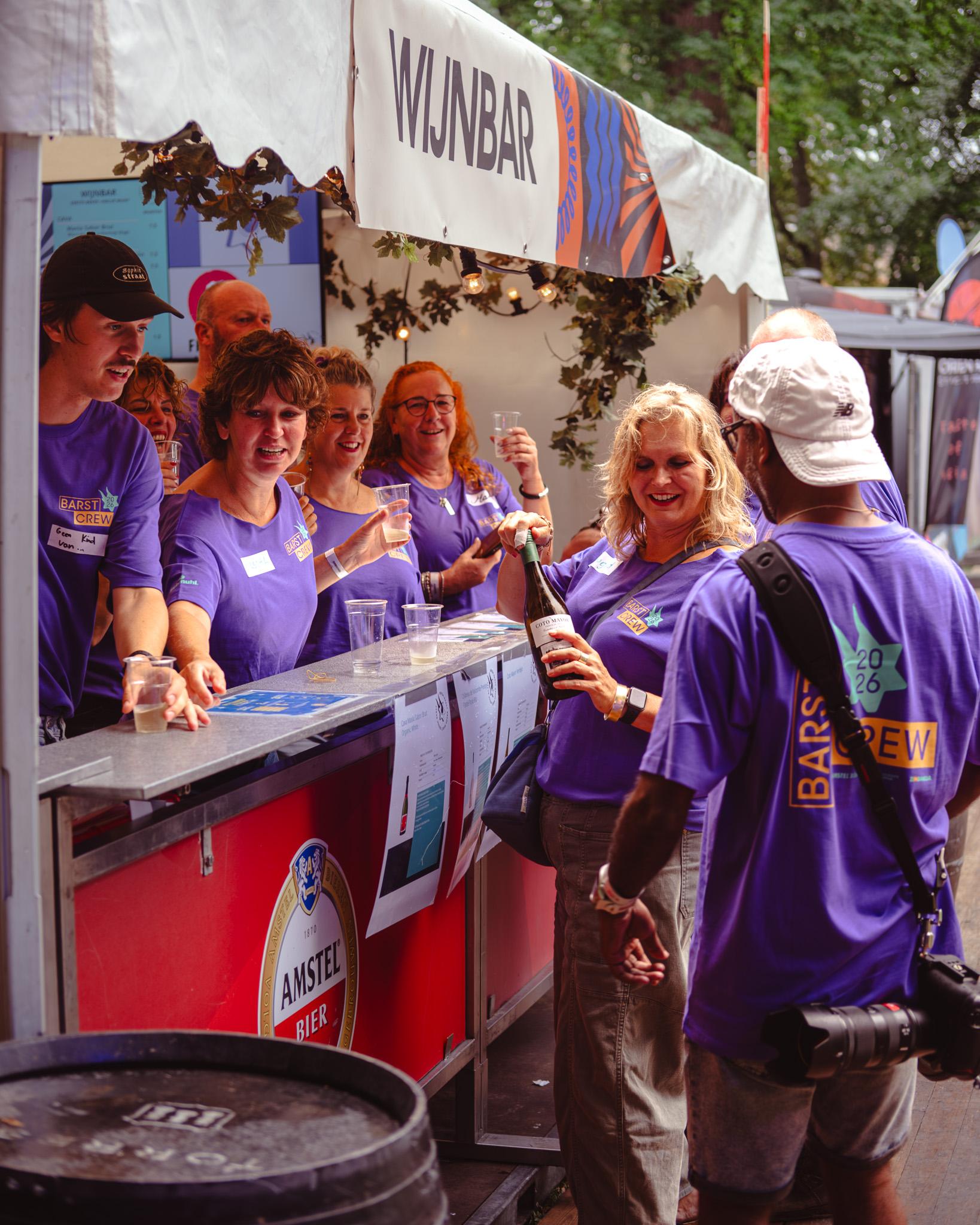 Barst Crew volunteers in purple t-shirts pouring wine and laughing together at the Wijnbar stall